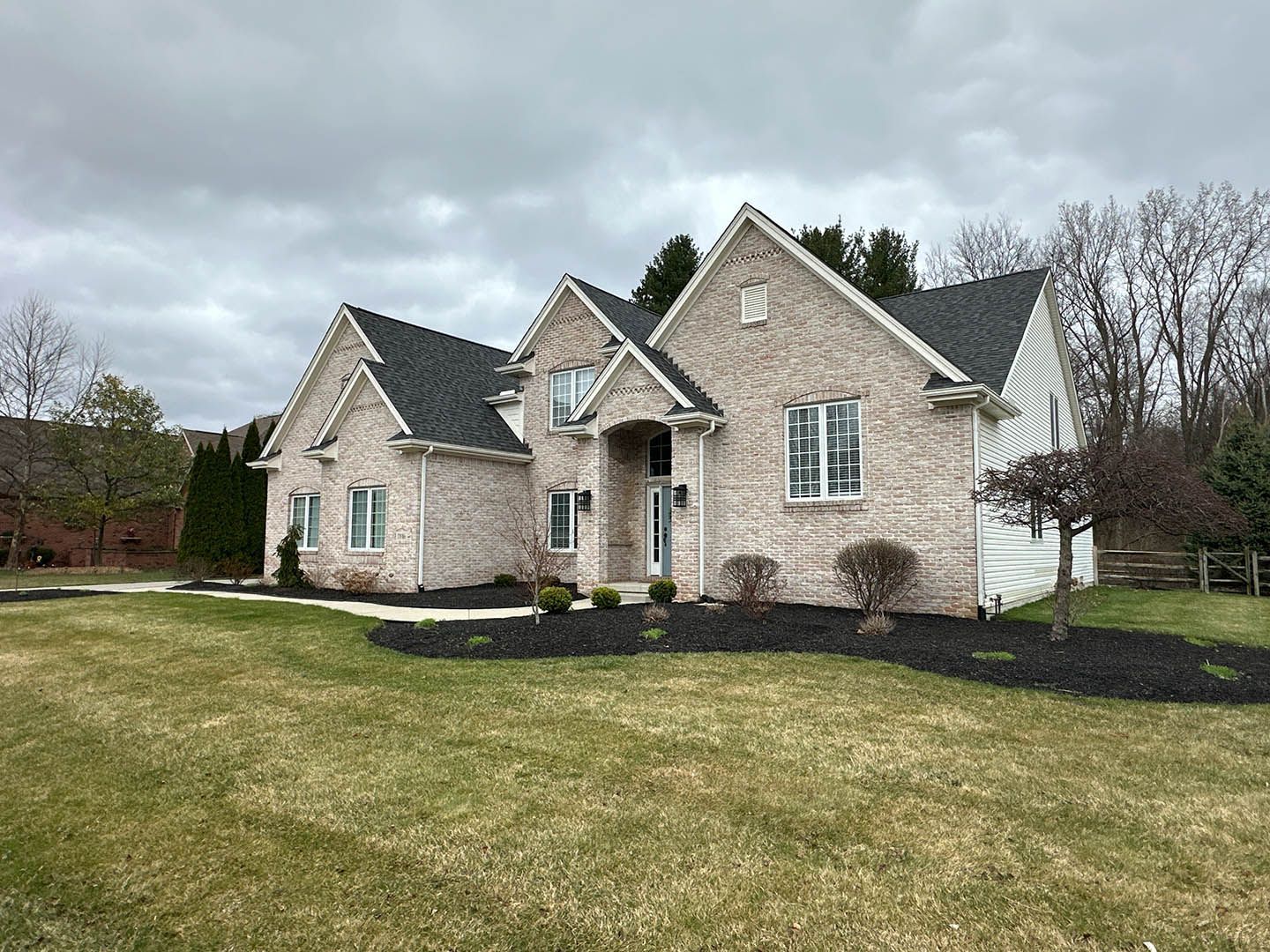 Suburban brick house with dark roof, white trim, and a manicured lawn under cloudy skies