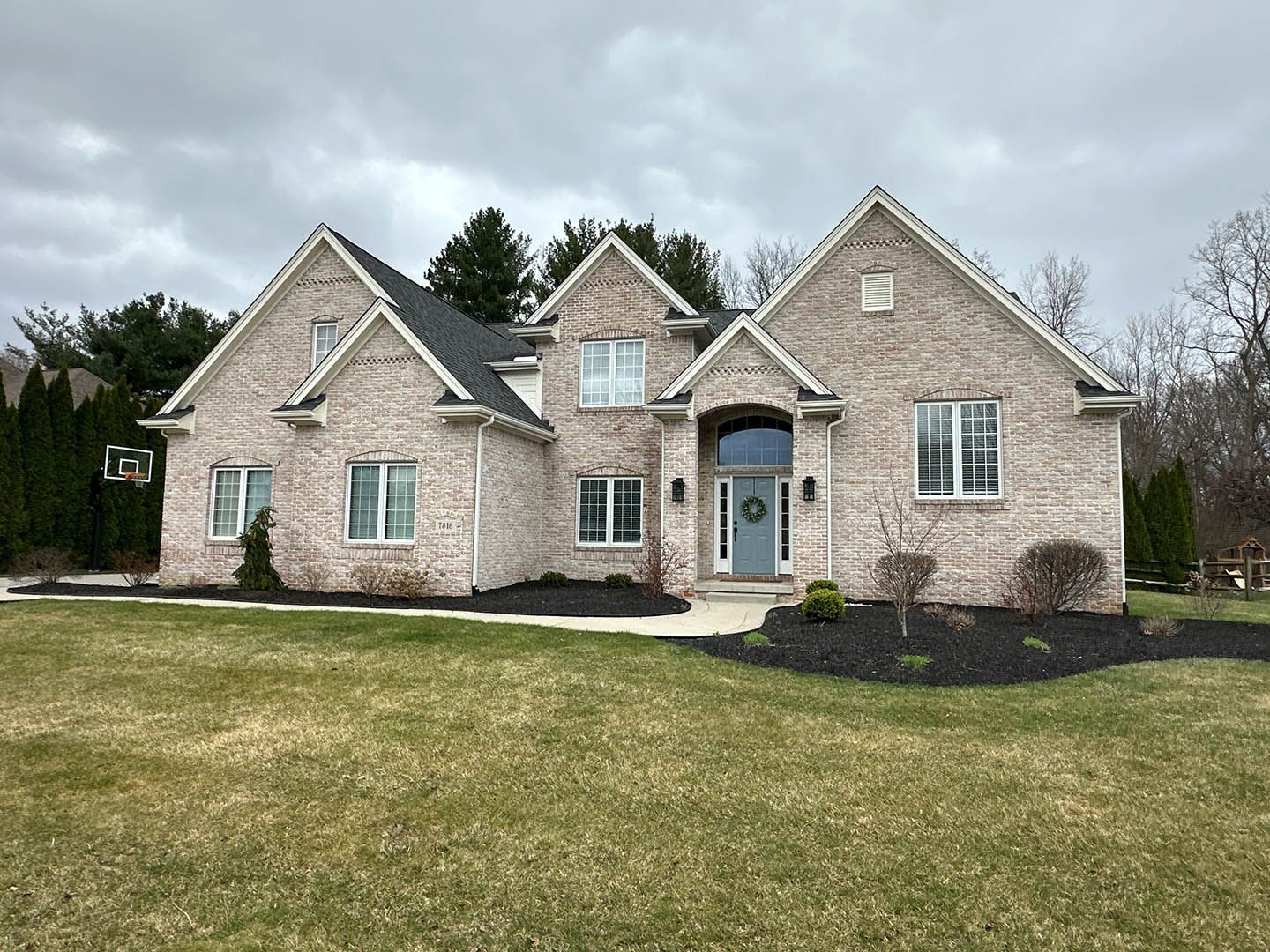 Large stone suburban house with gabled roof, front porch, and wide lawn under cloudy sky