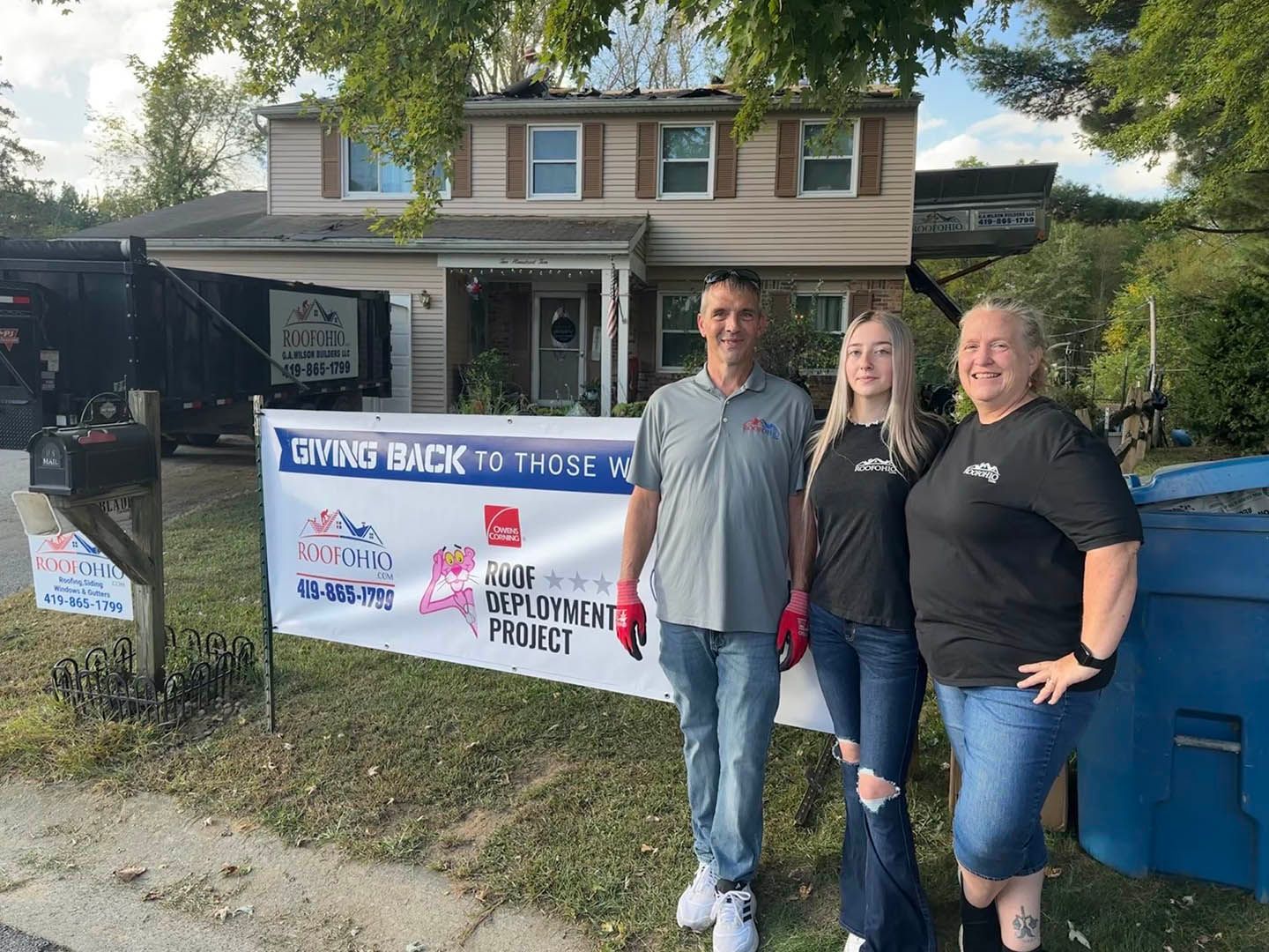 Three people posing beside a banner in front of a house at an outdoor event