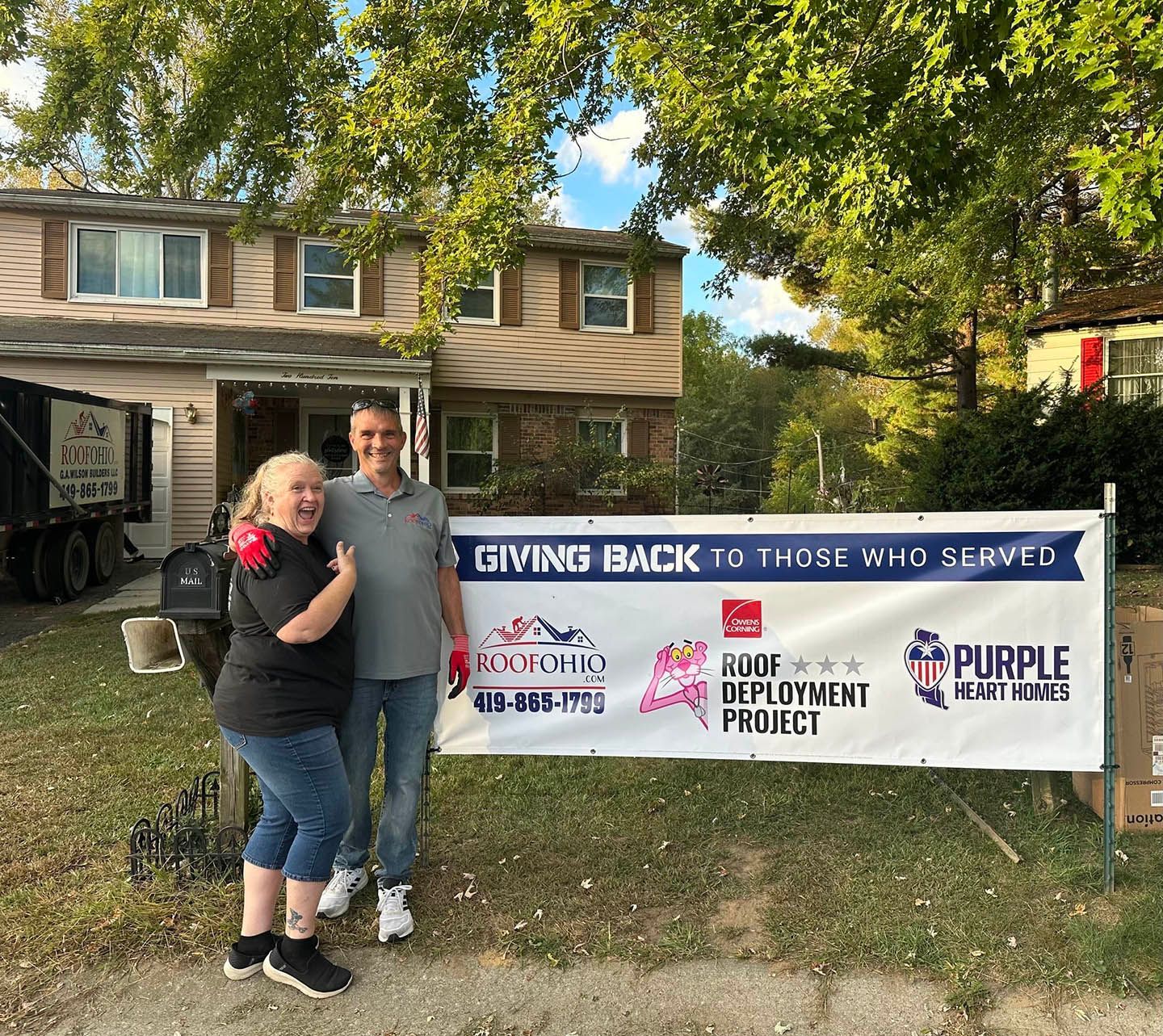Two people posing beside a “Giving Back to Those Who Served” banner in front of a house