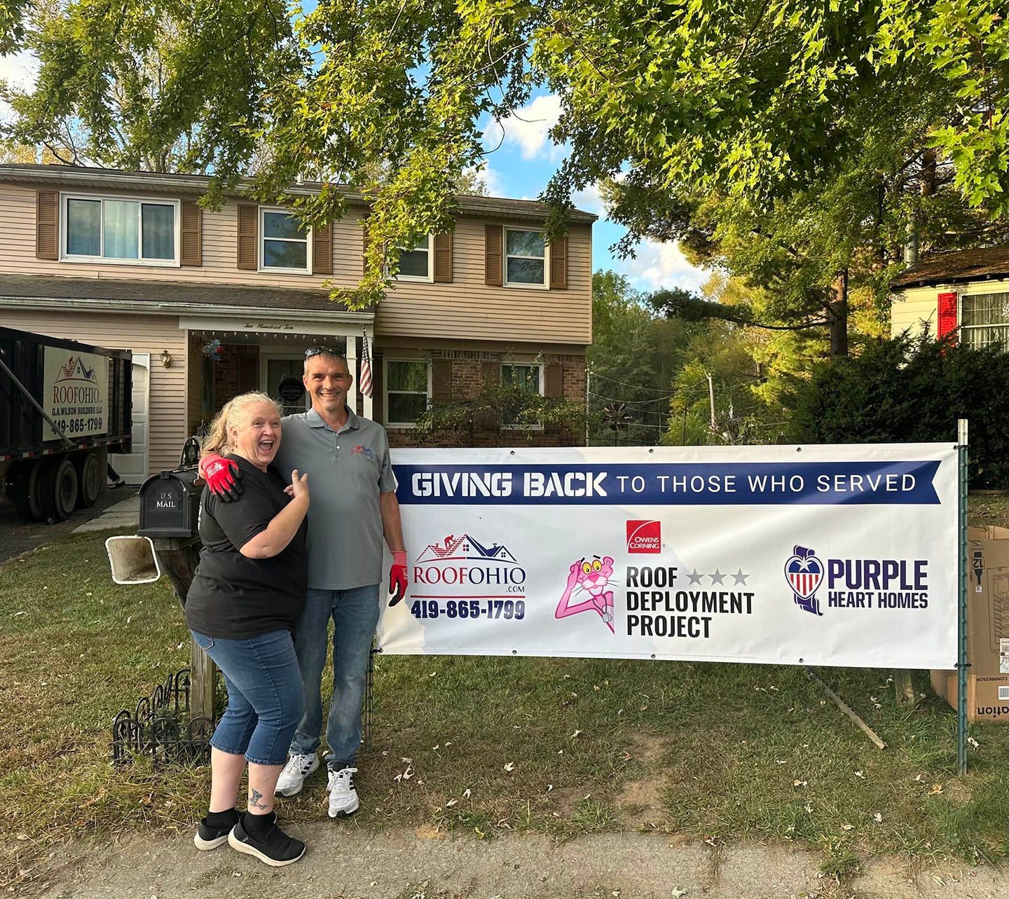 Two people pose beside a “Giving Back to Those Who Served” banner in front of a house.