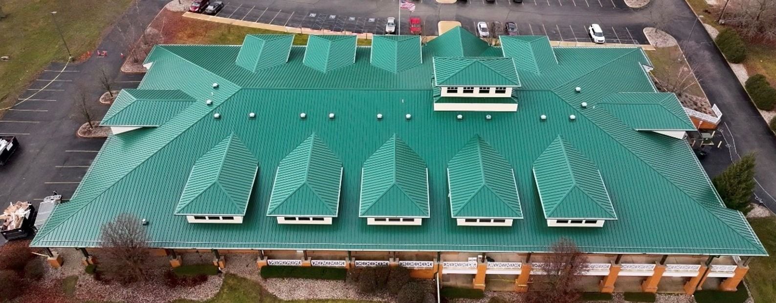 Aerial view of a building with a green metal roof and multiple skylights, surrounded by parking lots and trees.