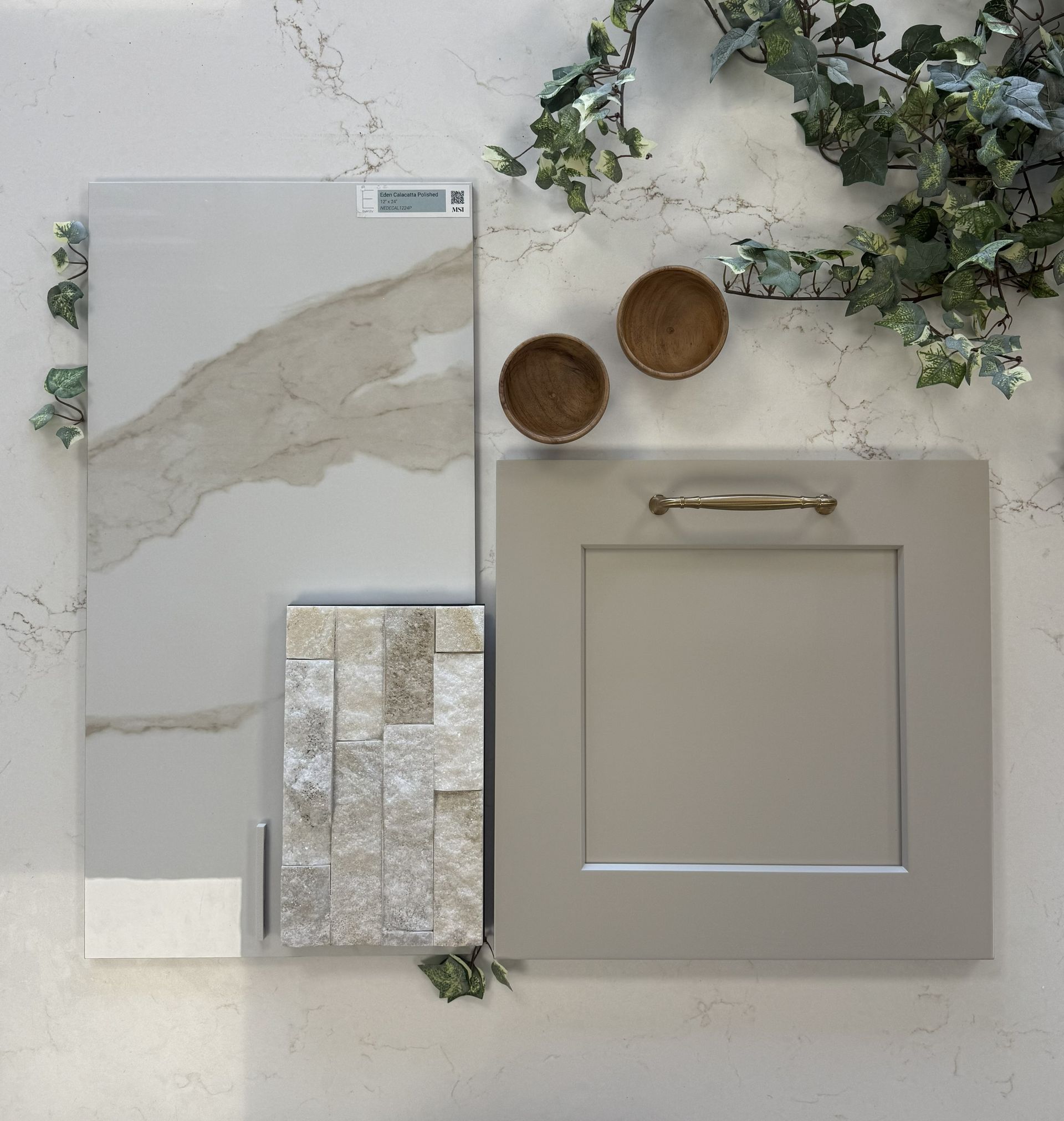 A design mood board with a marble slab, stone tile sample, cabinet door, brass hardware, and greenery on a white surface.