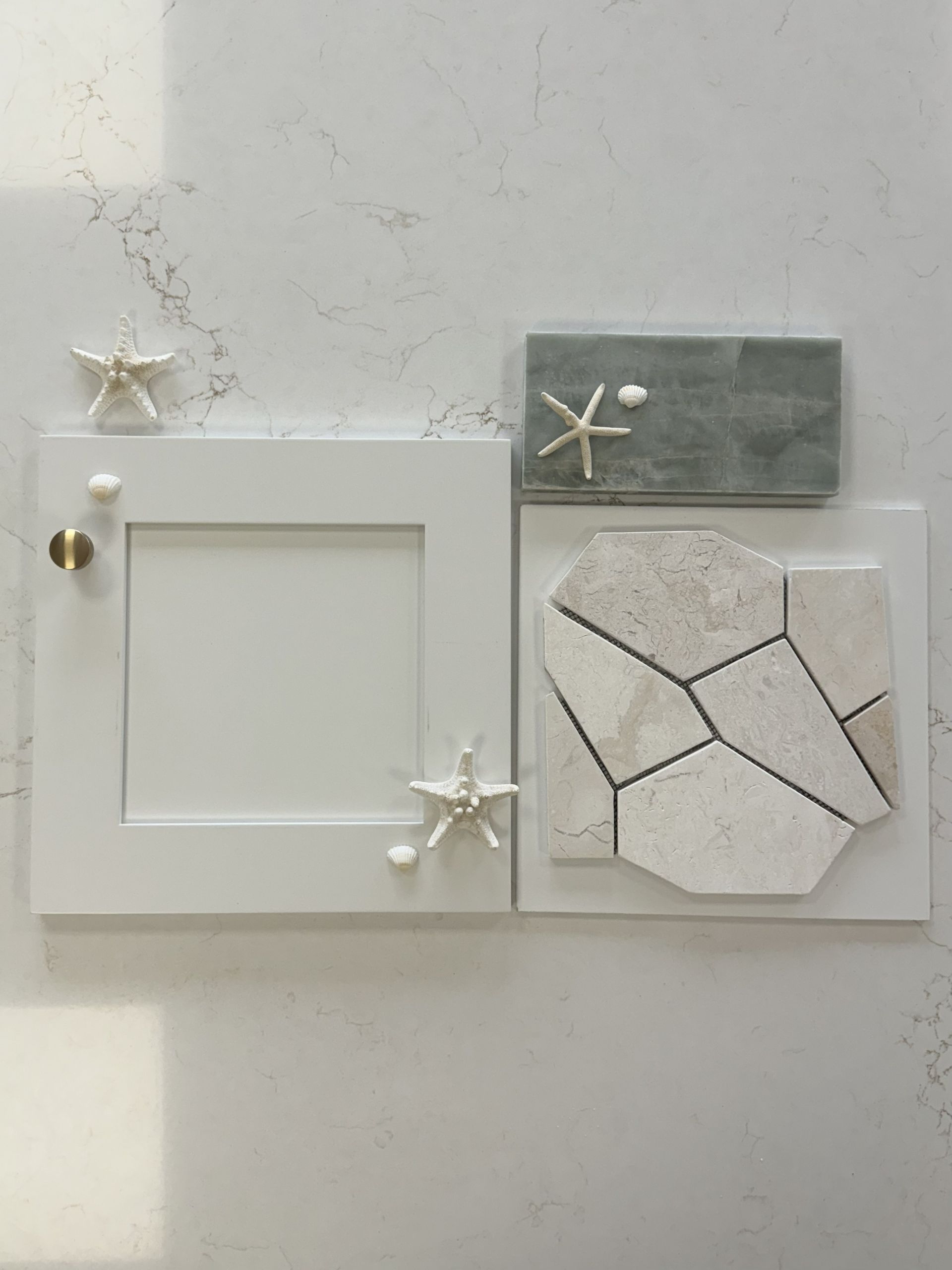 A flat-lay of a white cabinet door, a brass knob, a gray stone sample, and a geometric-patterned stone tile with seashells.