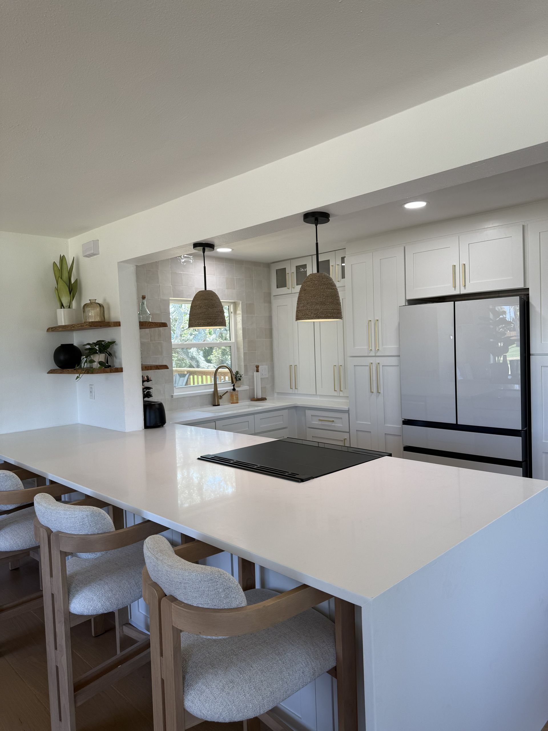 A bright kitchen featuring a large white island with woven bar stools, white cabinets, and two hanging textured pendants.