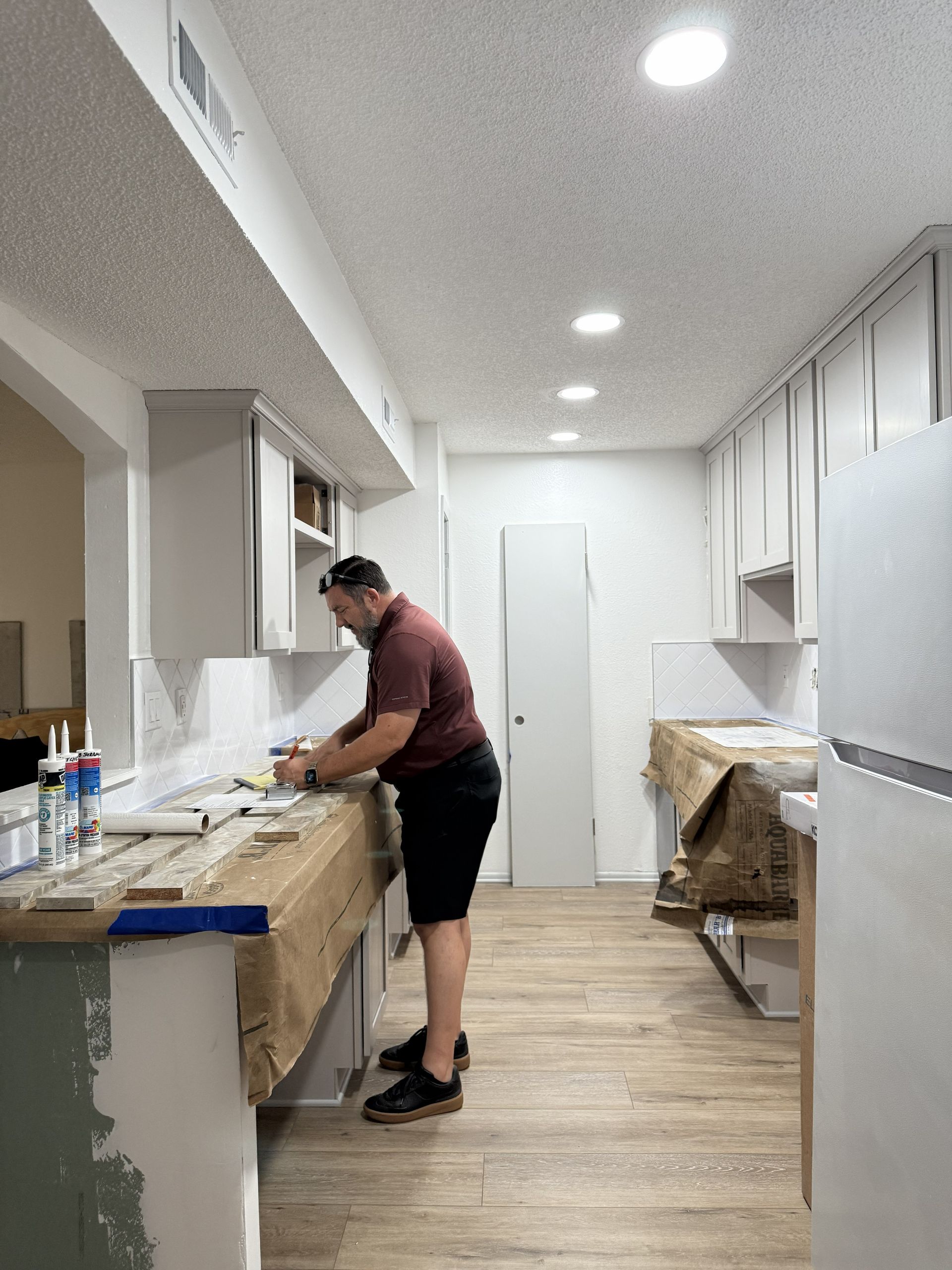 A person in a maroon shirt working on a counter in a kitchen undergoing renovations with white cabinets and wood floors.
