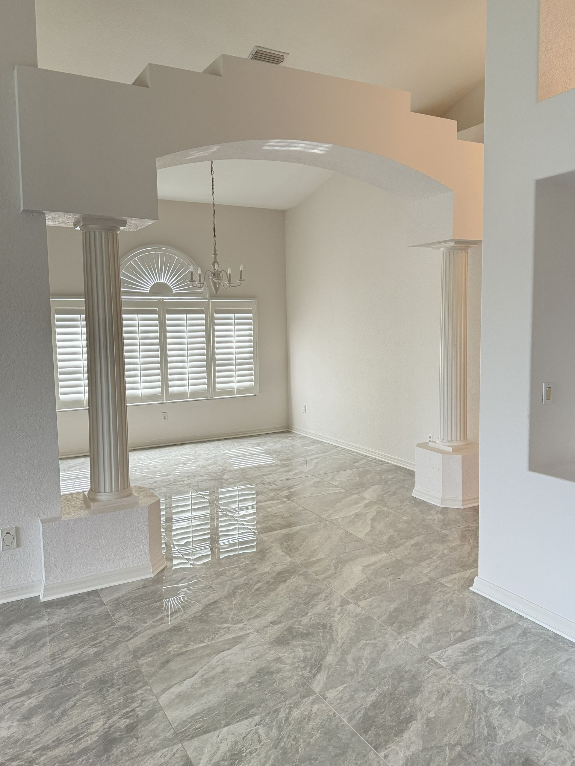 A bright, empty room with large grey marble-style floor tiles, white walls, and two decorative white pillars under an arch.