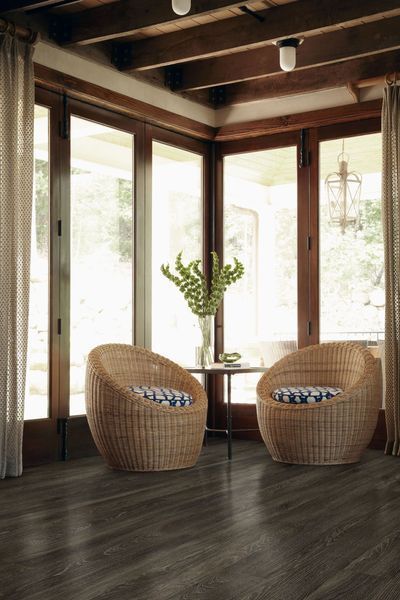 Two woven wicker chairs sit on a dark wooden floor by a corner window, with a small table and a plant between them.
