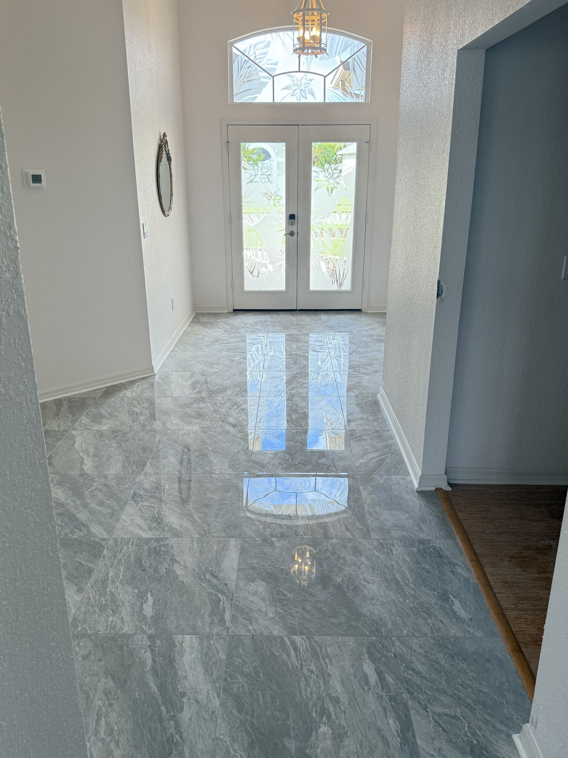 A bright entryway with polished grey marble-patterned tile flooring, leading to glass double doors and an arched transom.