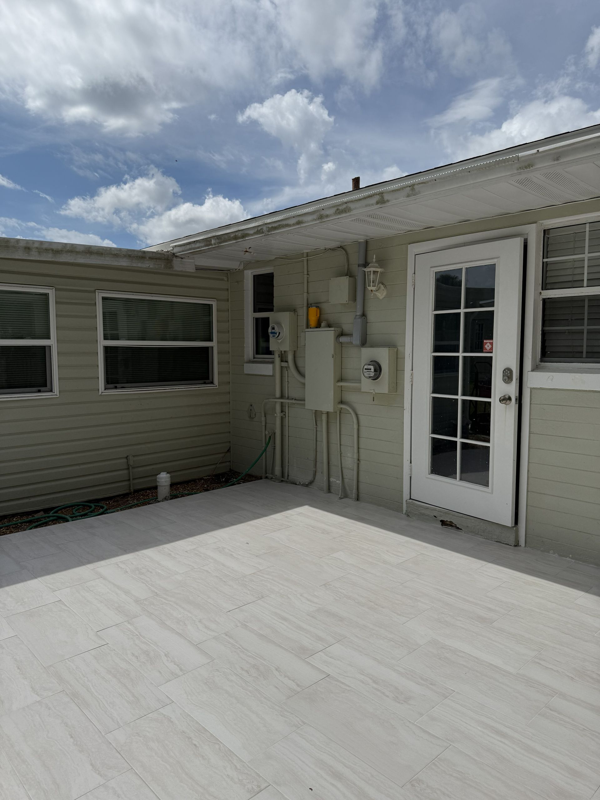 A patio area adjacent to a light-colored, single-story house with a white paneled door and several windows.