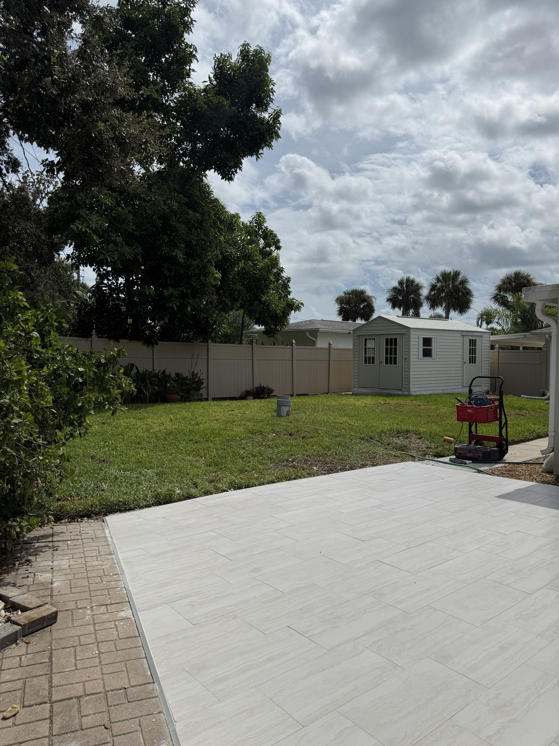 A backyard scene featuring a white concrete patio, a green grassy lawn, a small storage shed, and a privacy fence.