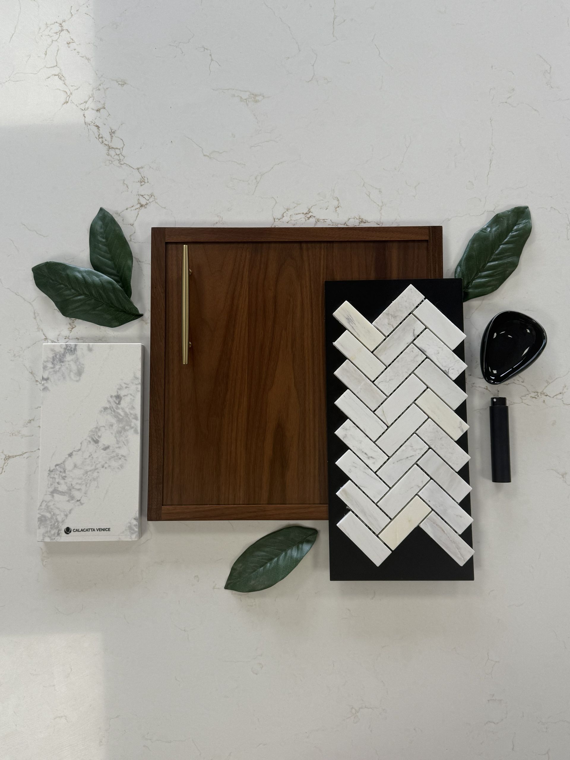 A design mood board with marble tile, a wood cabinet sample, gold hardware, and green leaves on a white quartz countertop.