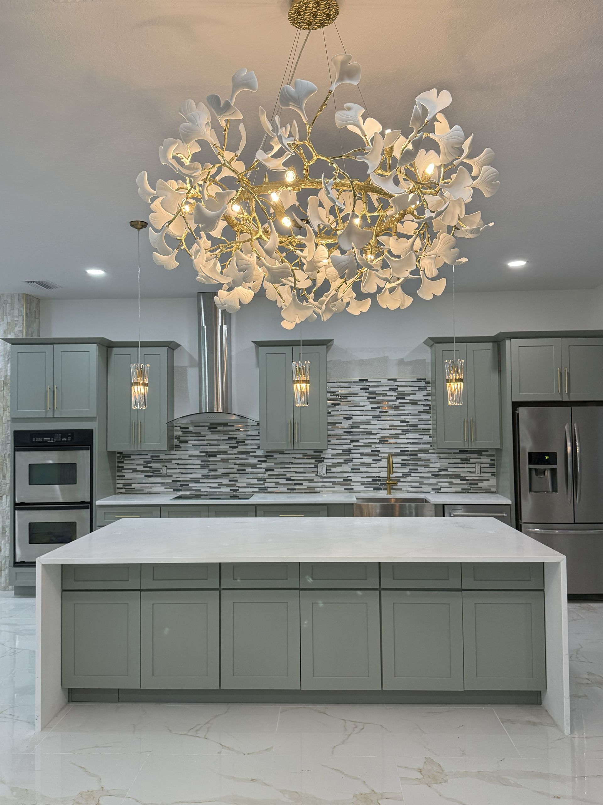 A kitchen with sage green cabinets, a white waterfall island, and a large, intricate floral-inspired gold chandelier.
