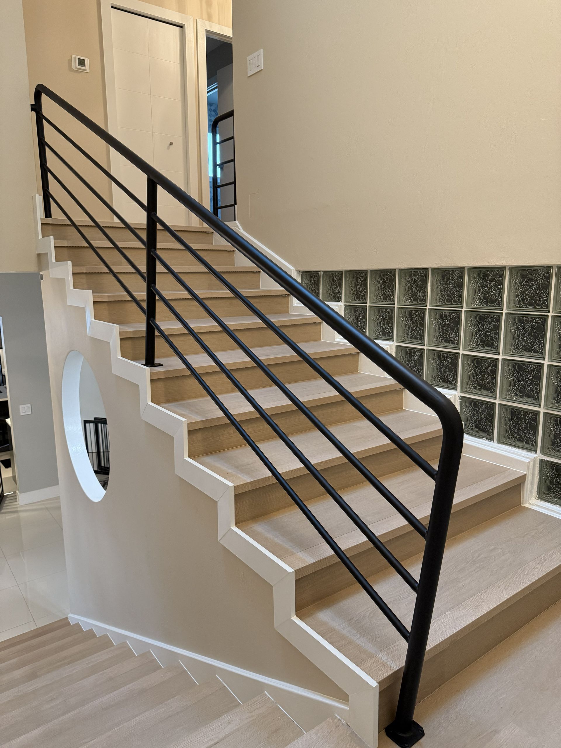 Interior staircase with light wood steps, white trim, a black metal horizontal railing, and a circular cutout in the wall.