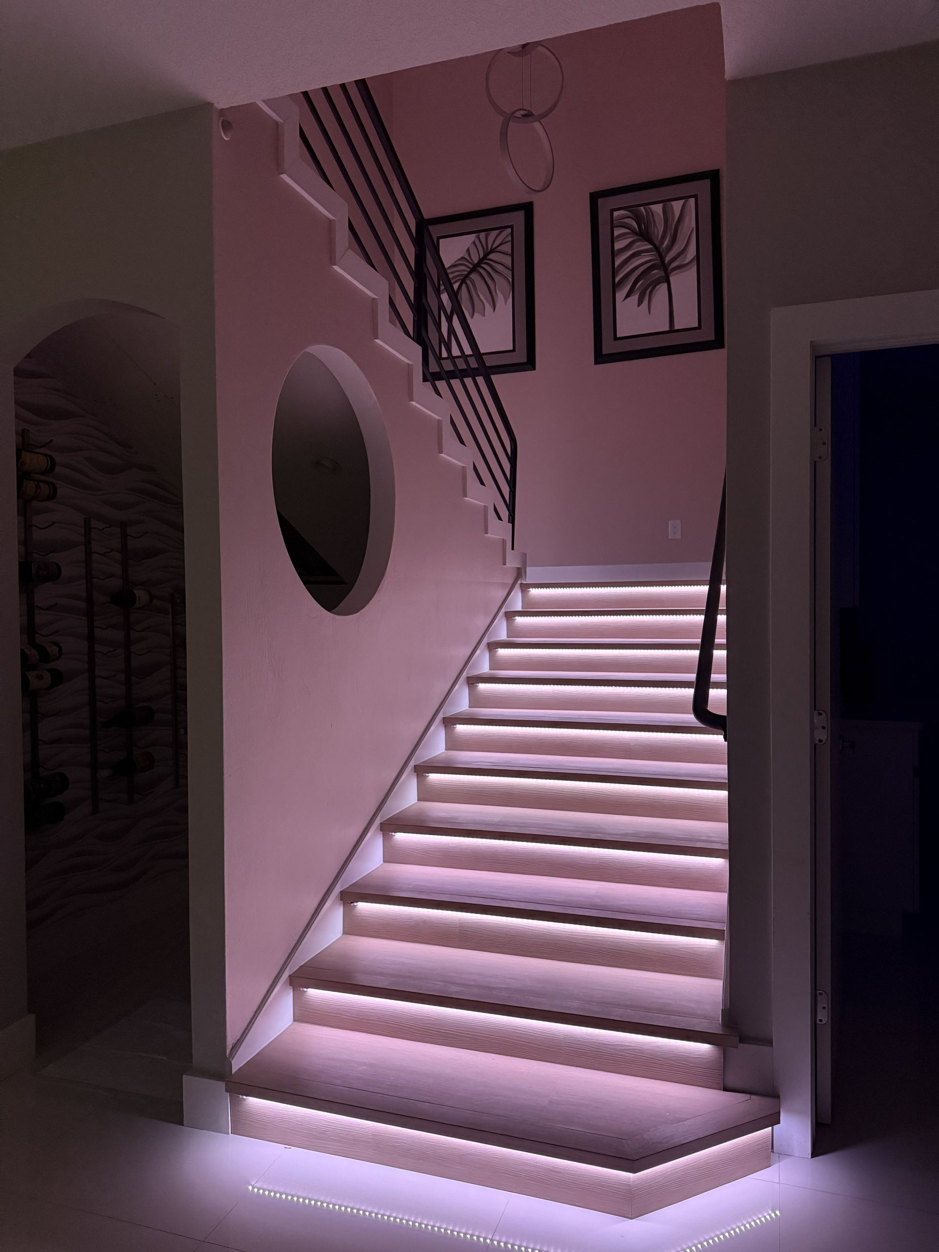 A set of carpeted stairs with illuminated steps, featuring a circular accent window and two framed prints on the wall.