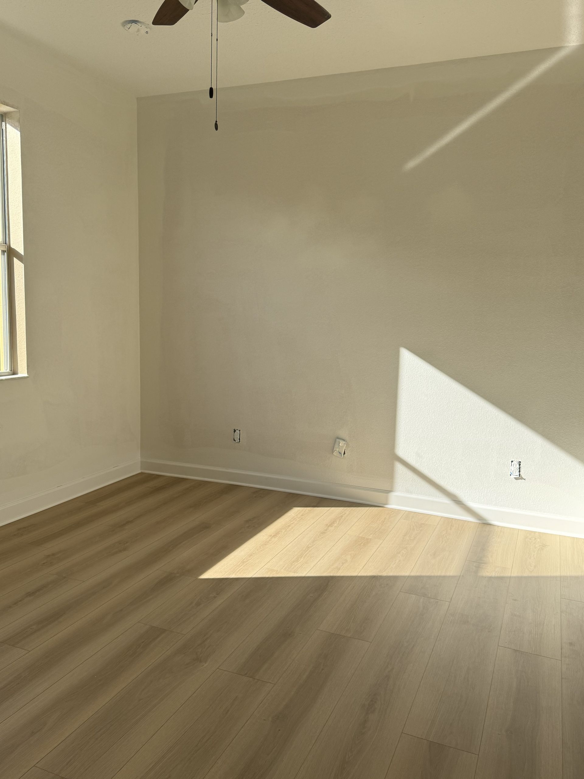 An empty room with light wood-look flooring, cream-colored walls, a ceiling fan, and a patch of sunlight on the floor.