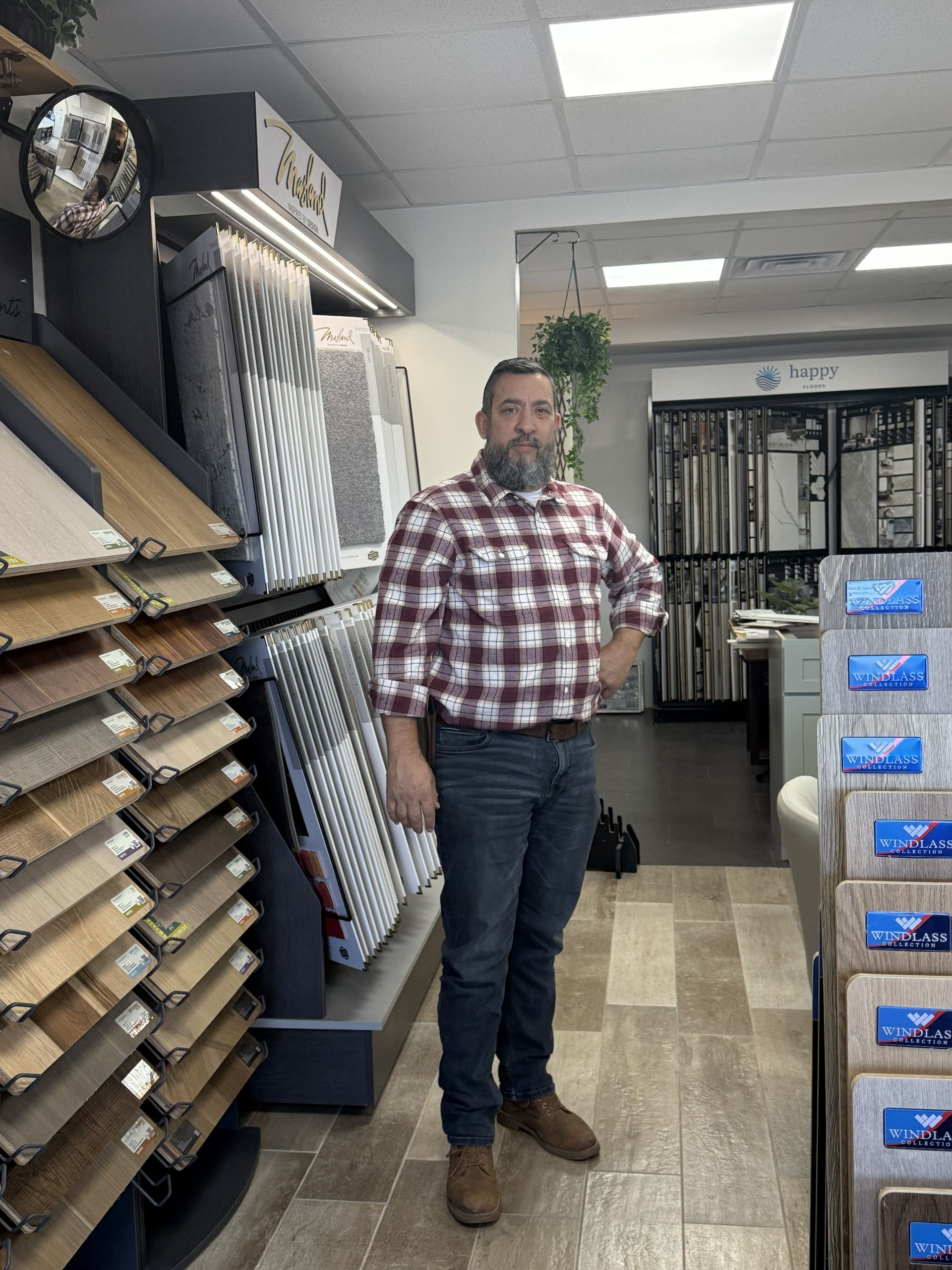 A man with a beard, wearing a plaid shirt and jeans, stands in a flooring showroom aisle surrounded by product samples.