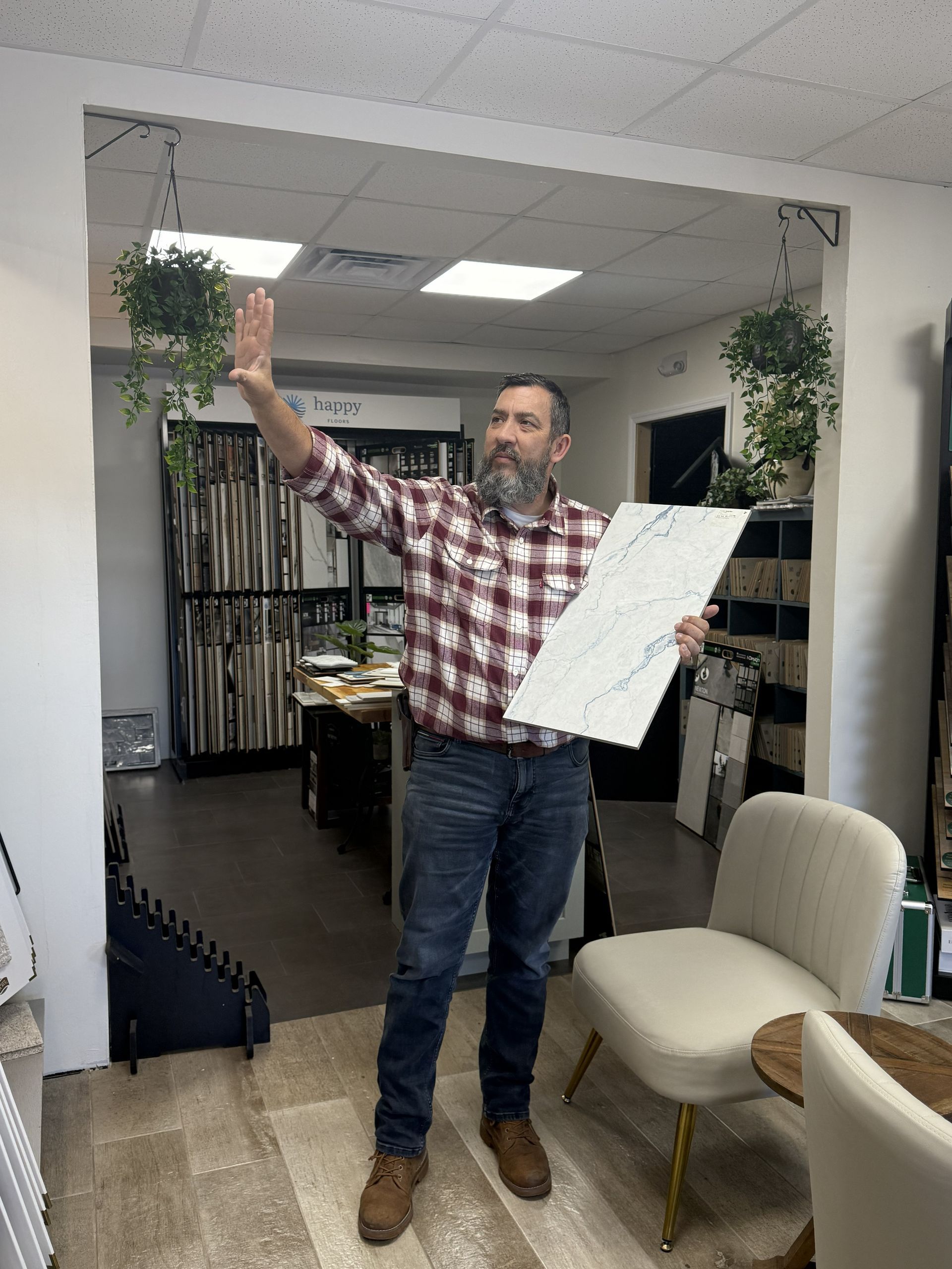 A person with a beard in a plaid shirt stands in a flooring showroom, gesturing and holding a white sample tile.