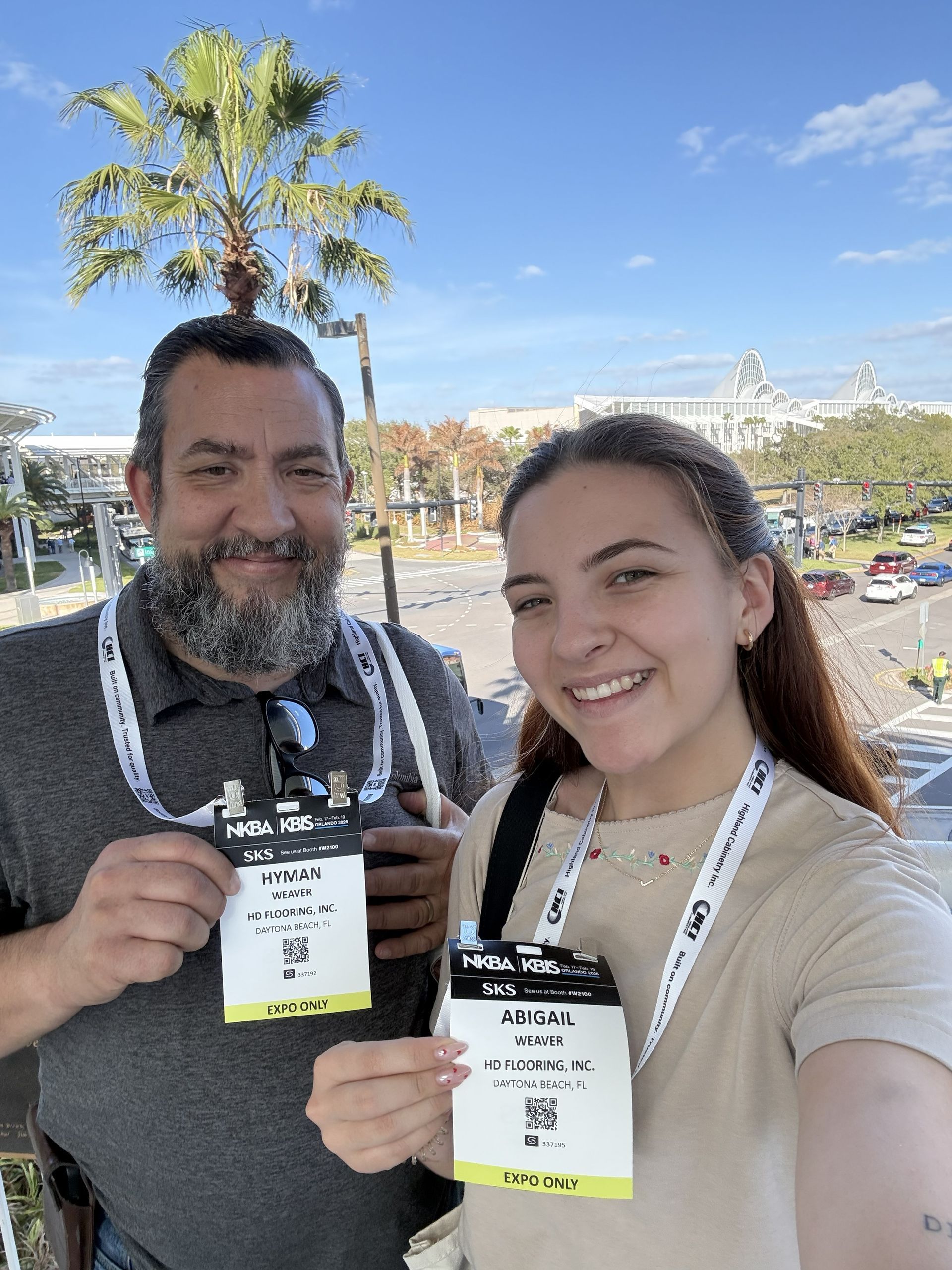 Two people smile while holding up event badges in front of a palm tree and a sunny, outdoor parking lot.