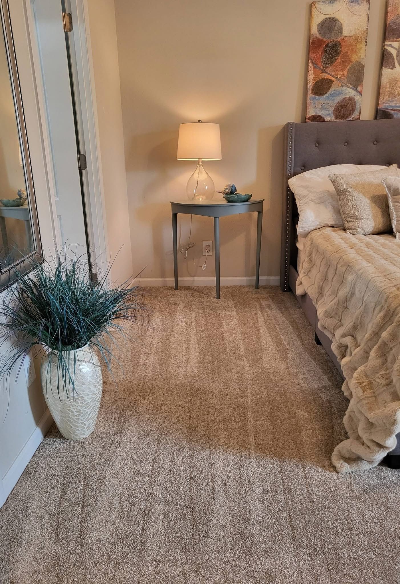 A bedroom corner with a gray tufted headboard, a round side table with a lamp, and a large vase of greenery on the floor.