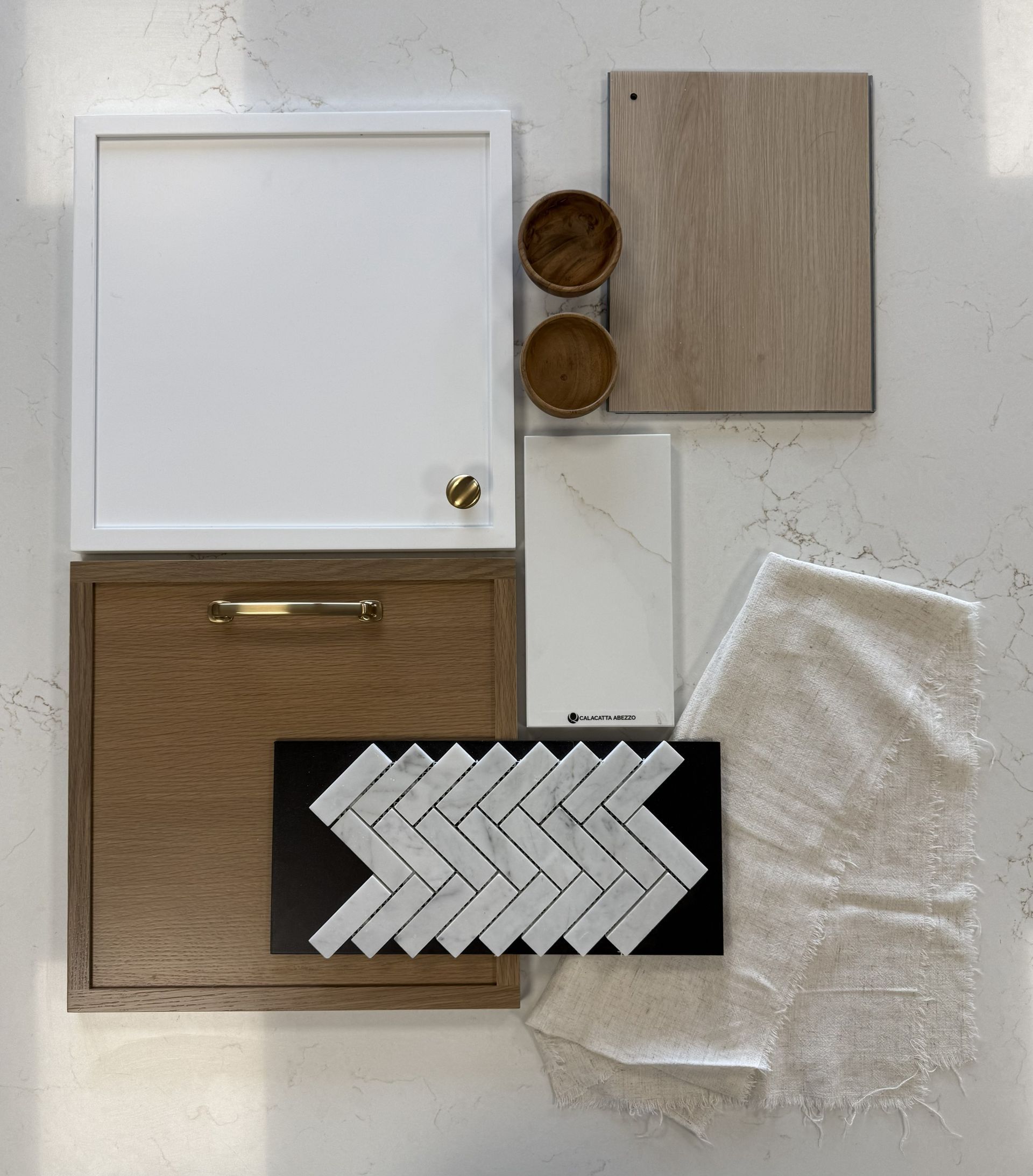 A flat-lay design board featuring wood, white cabinetry, brass hardware, marble herringbone tile, and textured fabric.
