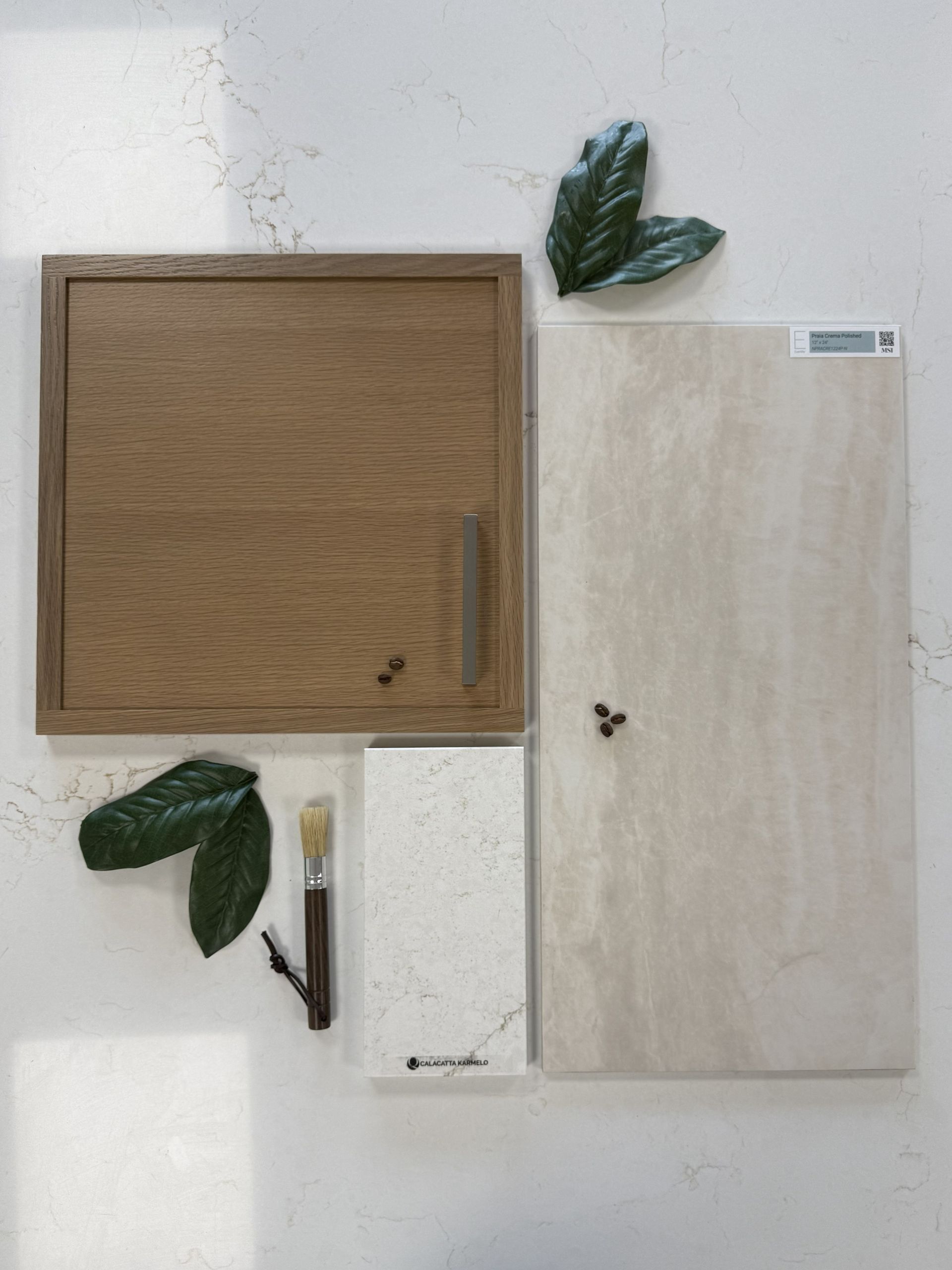 A flat-lay of interior design samples: wood cabinetry, stone tiles, a small brush, and leaves on a white background.