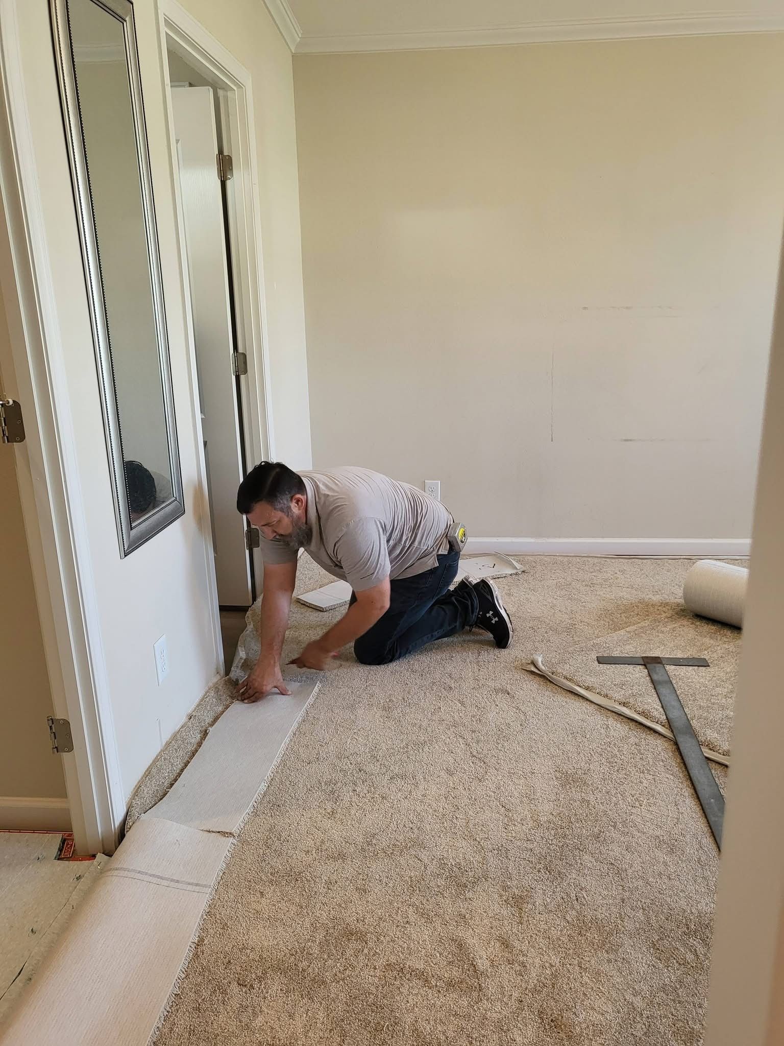 A professional kneeling on a light-colored carpet, using a tool to install or trim new carpeting near a door frame.
