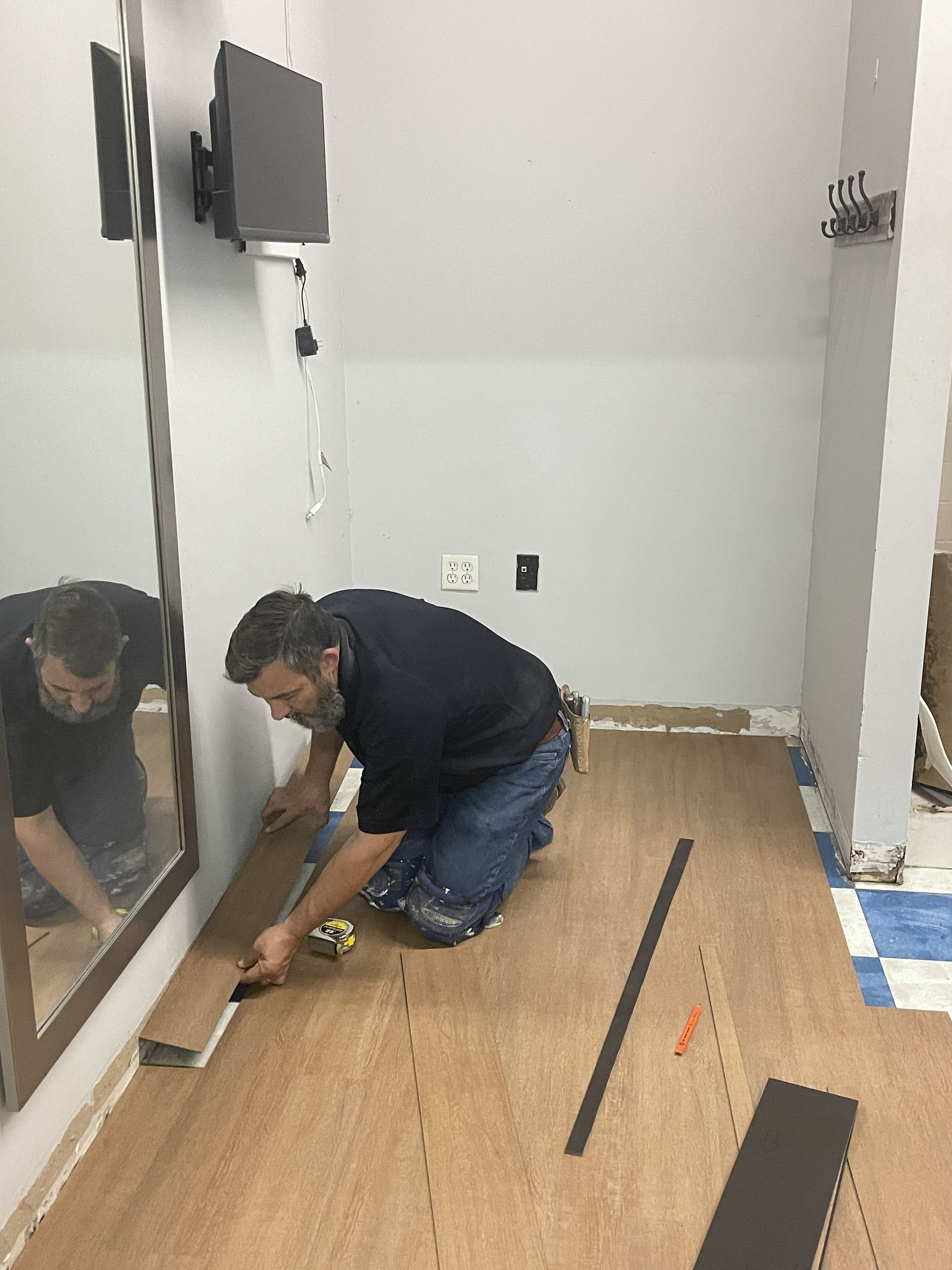 A person in a dark shirt and jeans kneels on a floor to install light brown wood-look planks in a bright room.