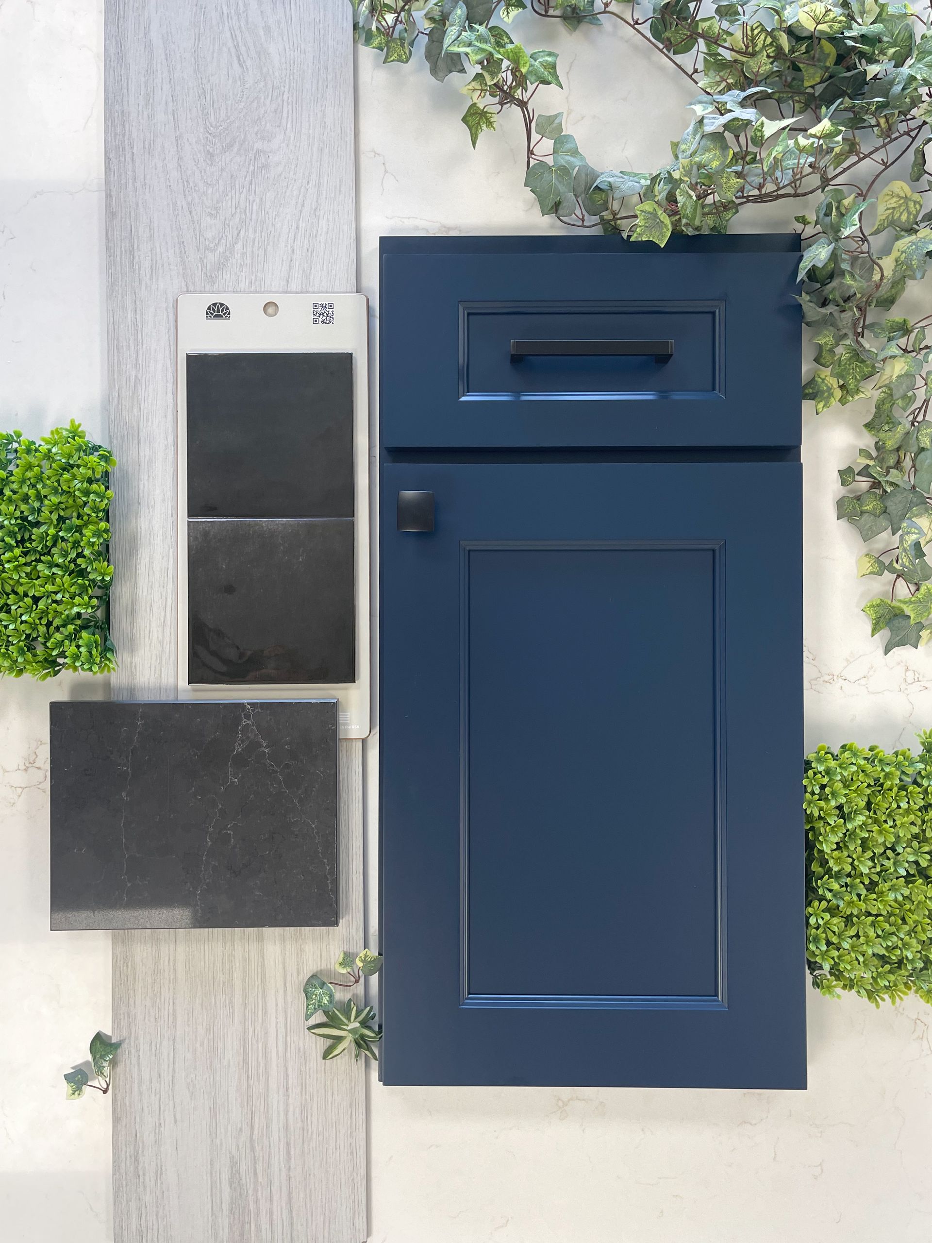 A sample board showing a navy blue cabinet door next to light wood flooring, a black stone slab, and decorative greenery.