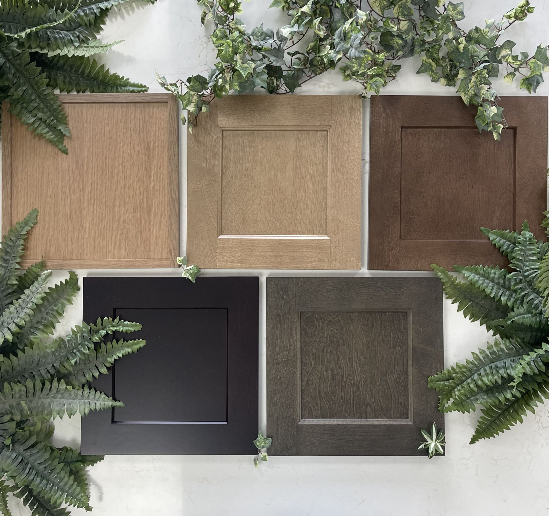 Five square cabinet door samples in varying wood tones and black, arranged in a grid surrounded by green foliage.