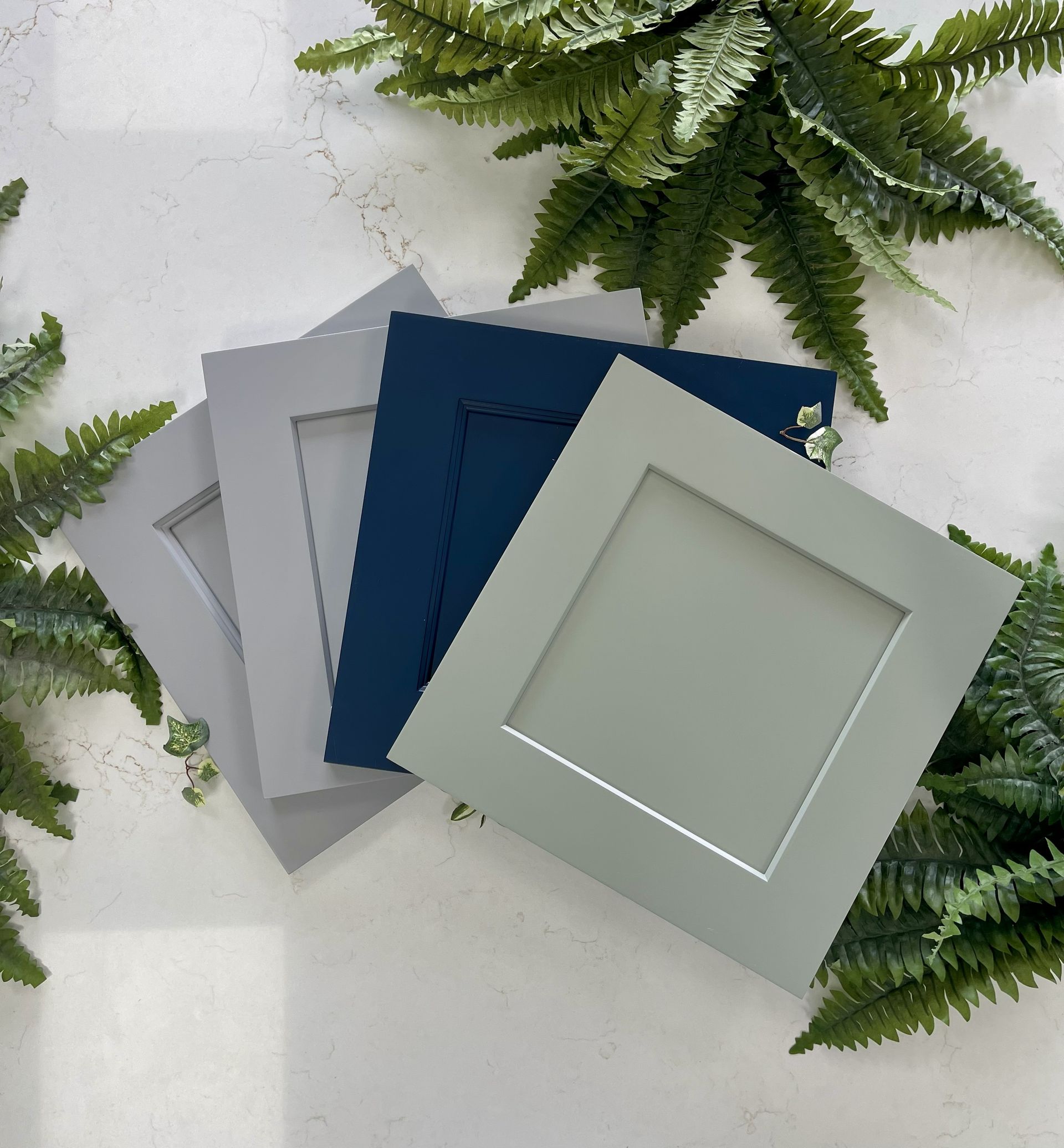 A display of four square-shaped cabinet door samples in varying shades of gray, dark blue, and light green on a white base.