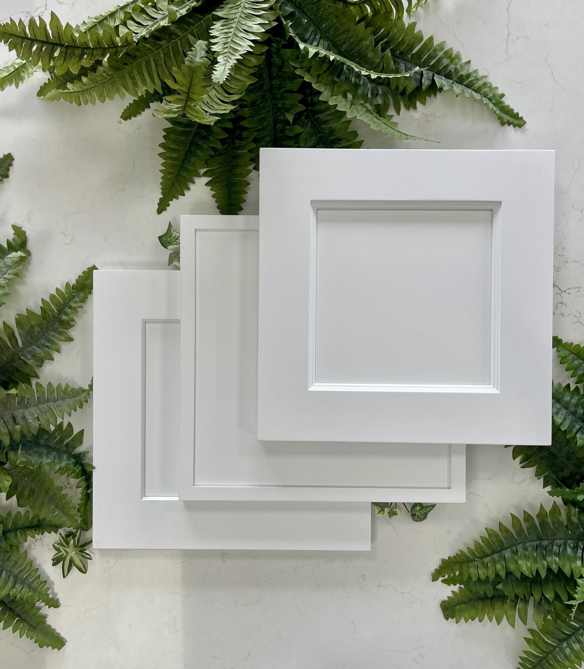 Three square white cabinet door samples layered over each other, set against a white background with green fern accents.