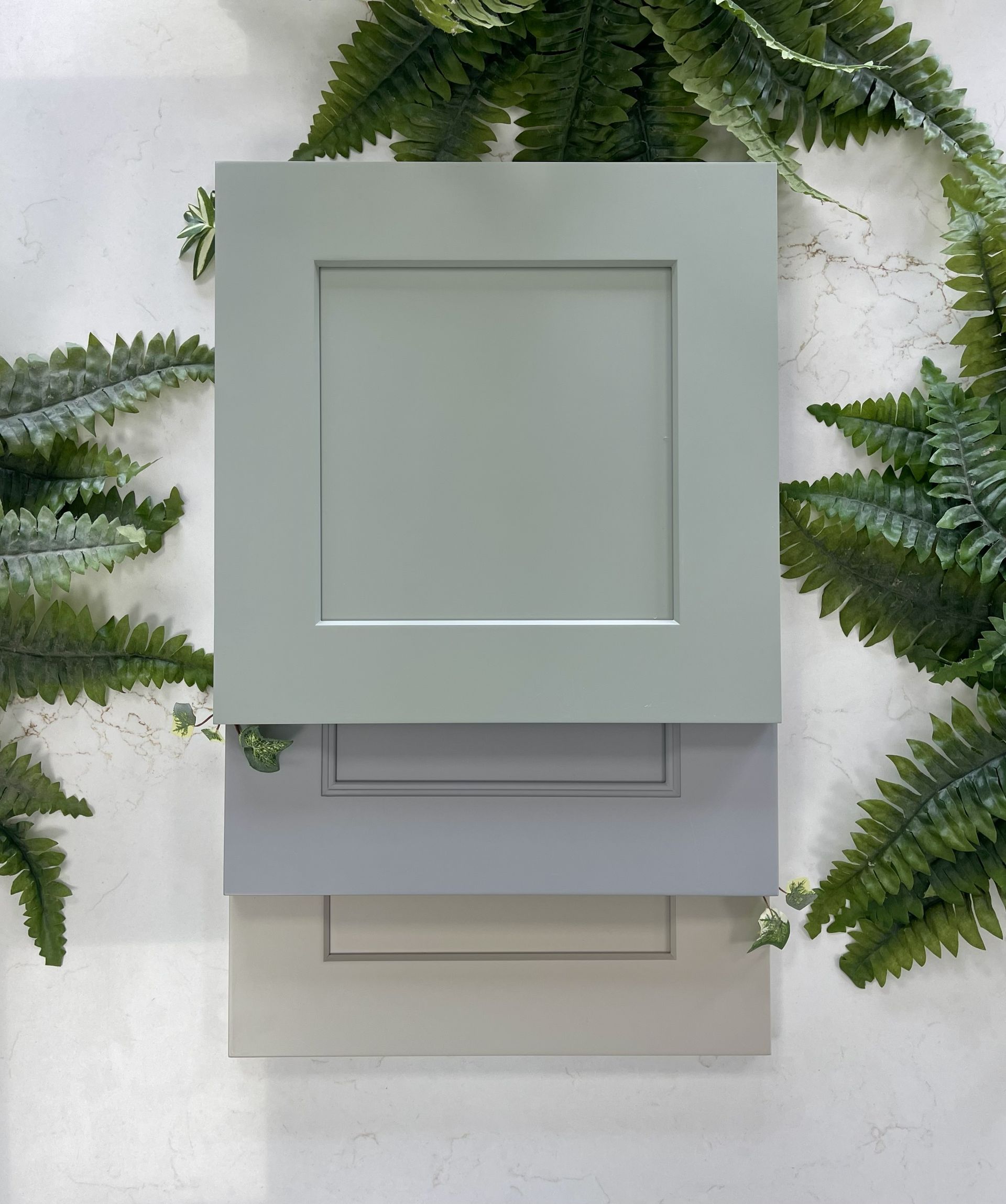 Three shaker-style cabinet door samples in sage green, grey, and beige, displayed against a white wall with green ferns.