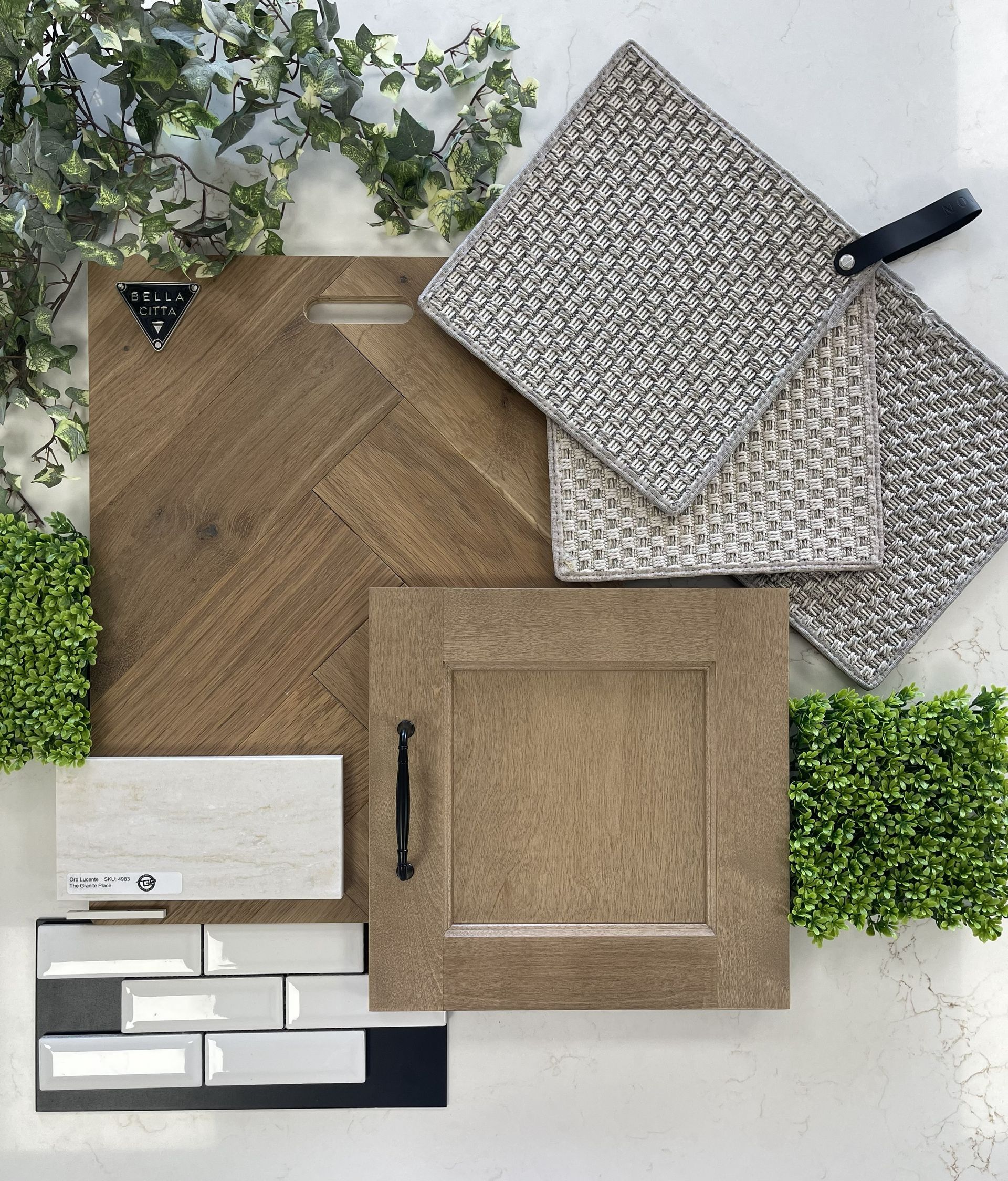 A flat-lay of interior design materials: wood flooring, a cabinet door sample, neutral tiles, textured fabric, and greenery.