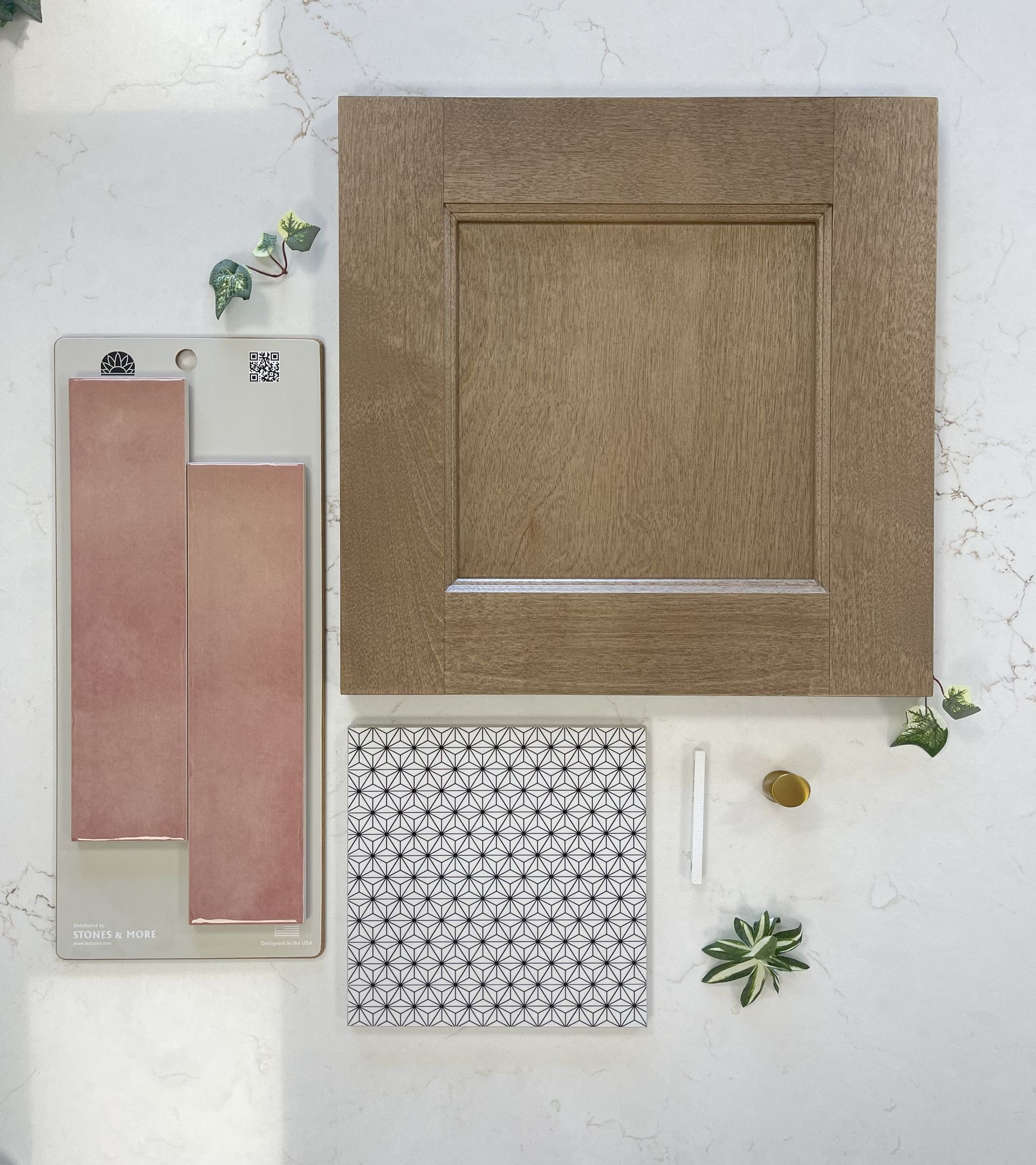 A mood board with a wood cabinet sample, two dusty rose tiles, patterned fabric, a white stick, and small green sprigs.