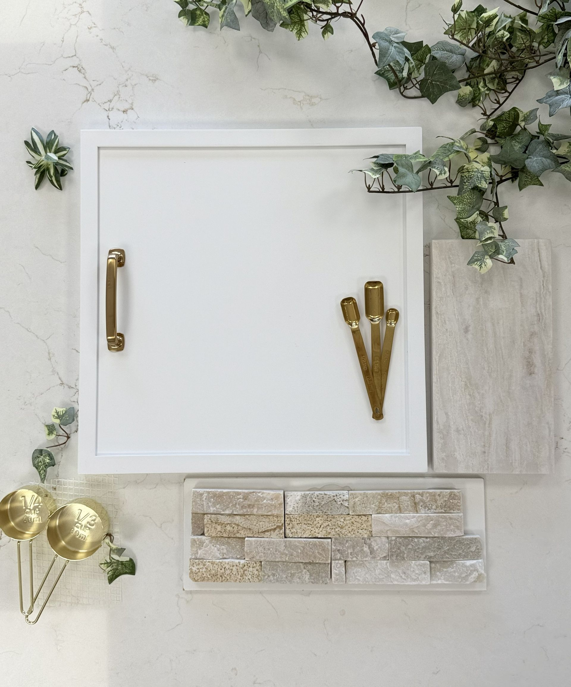 A design mood board featuring a white cabinet sample, gold hardware, textured stone tiles, and decorative greenery.