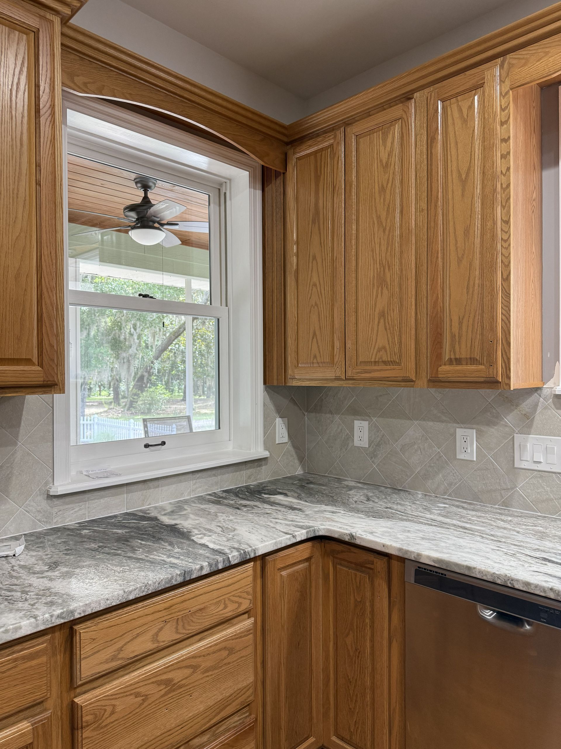 A kitchen corner with oak wood cabinets, a window looking outside, and white granite countertops with grey veining.