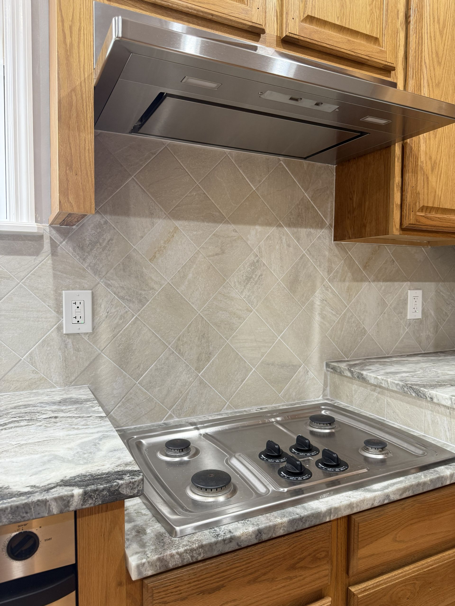 A kitchen stovetop with a stainless steel range hood, granite countertops, and a diagonal tile backsplash.
