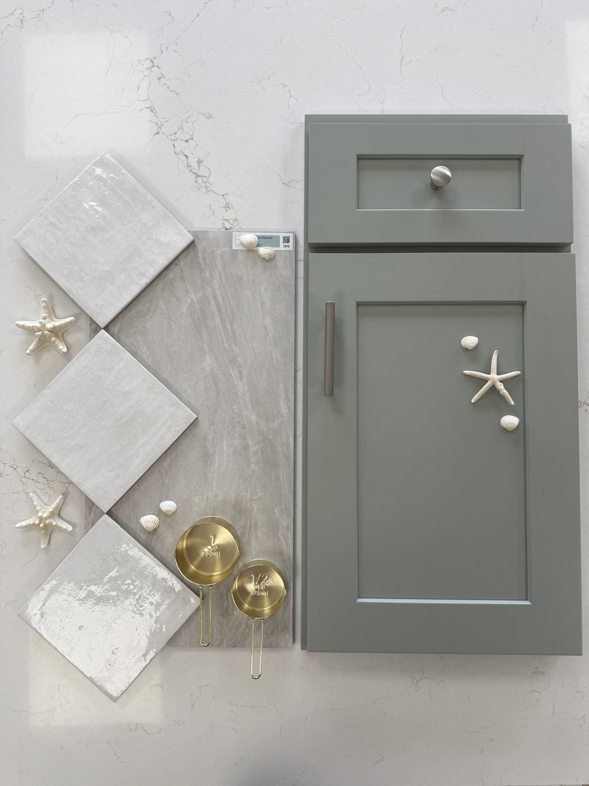 A flat lay shows a gray cabinet door, three square white tiles, stone samples, and decorative starfish on a marble surface.