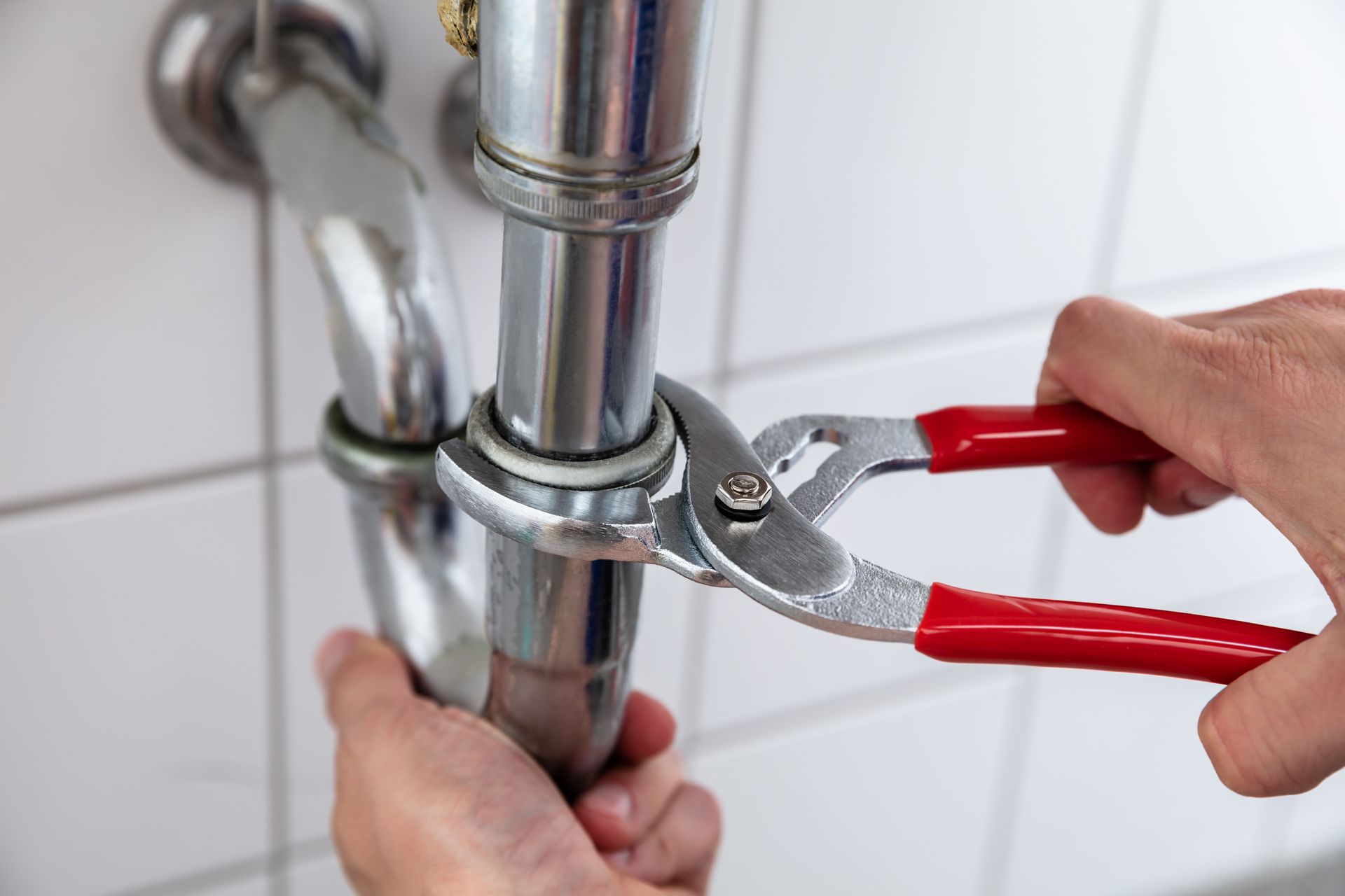 Hands using red-handled pliers to tighten a chrome pipe under a sink