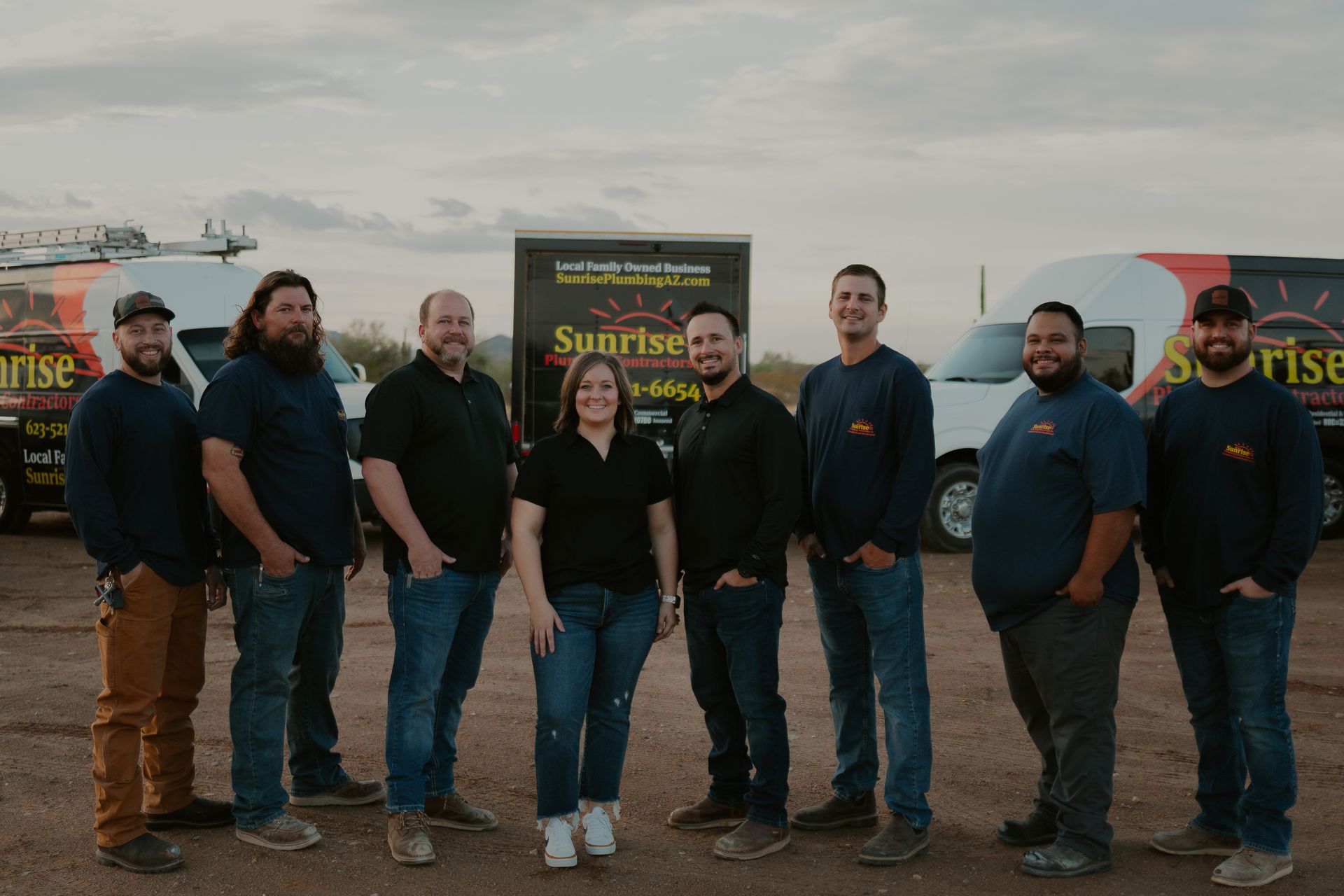 Group of 8 people posing outside with work vans. They are wearing work clothes.