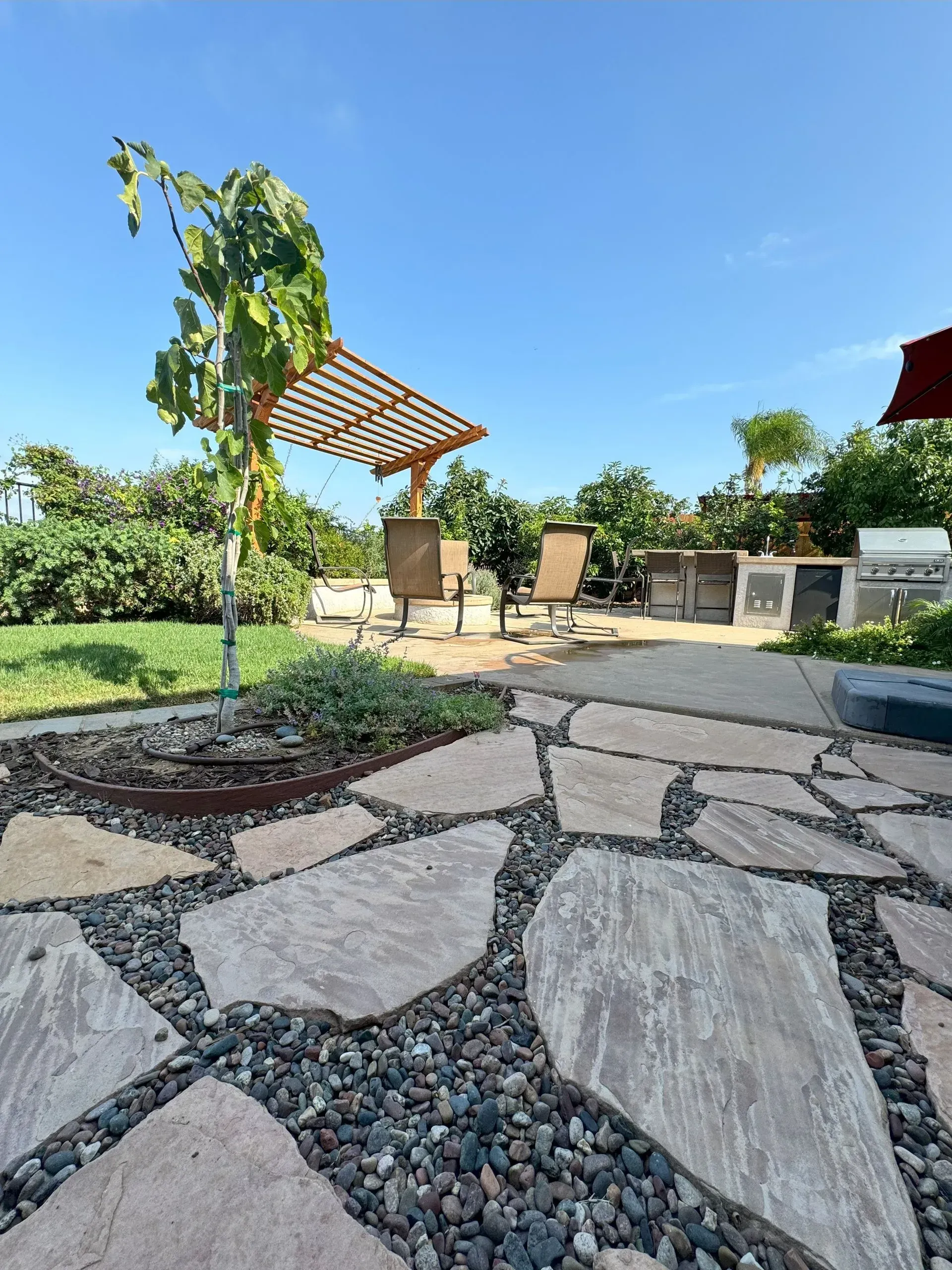 Patio with flagstone path, seating area under pergola, and lush greenery against a blue sky.