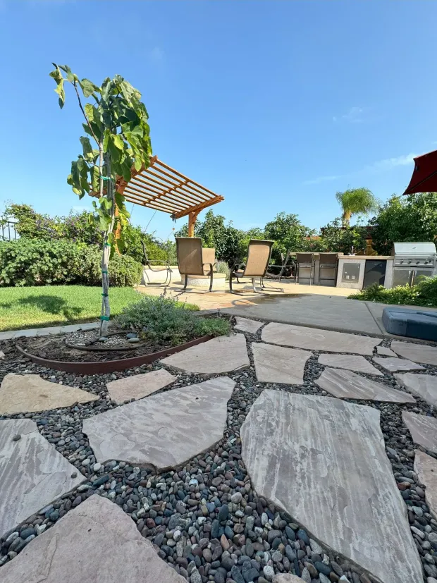 Patio with flagstone path, seating area under pergola, and lush greenery against a blue sky.