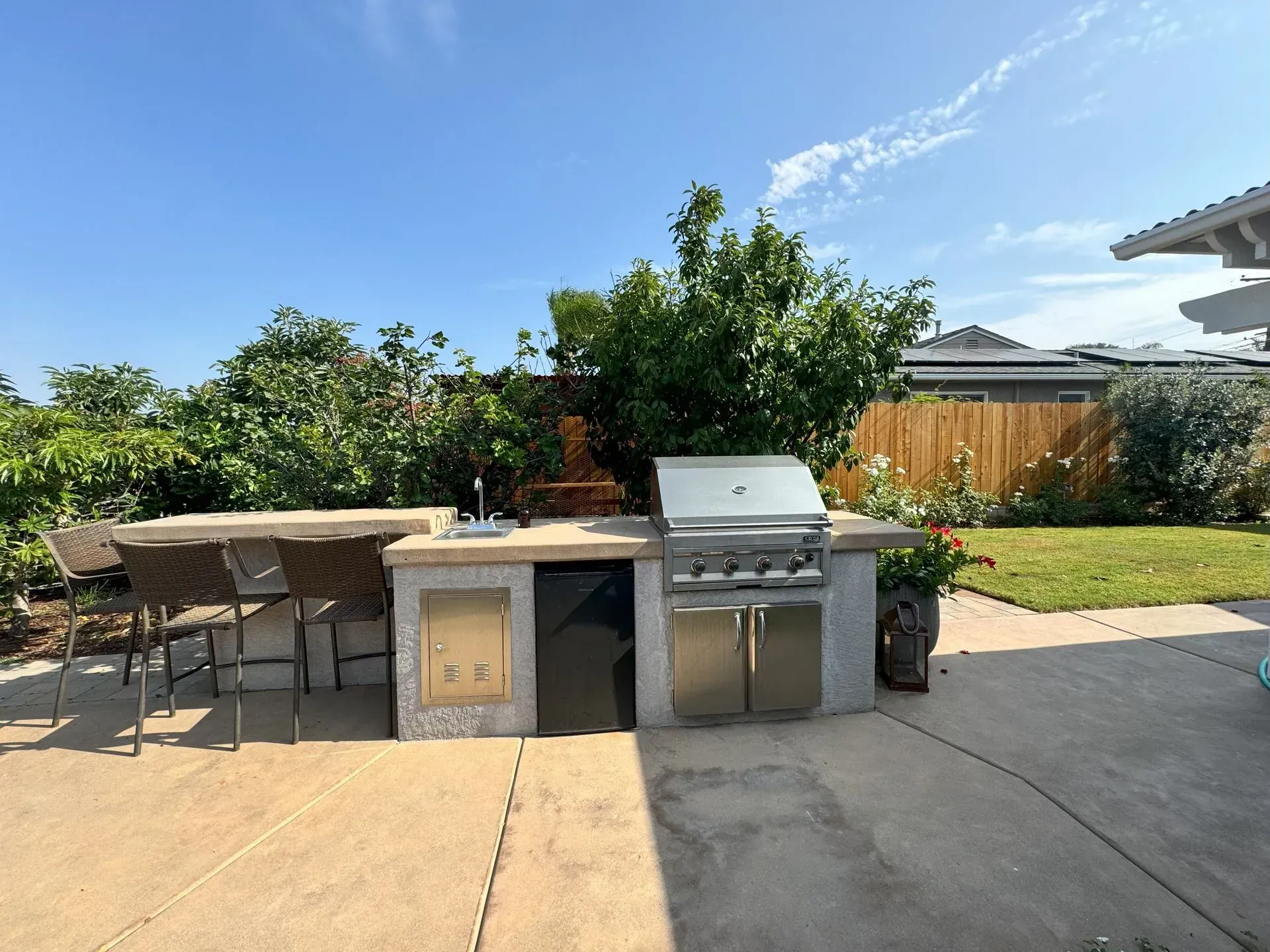 Outdoor kitchen with grill, counter, bar stools, and lush greenery in a sunny backyard.