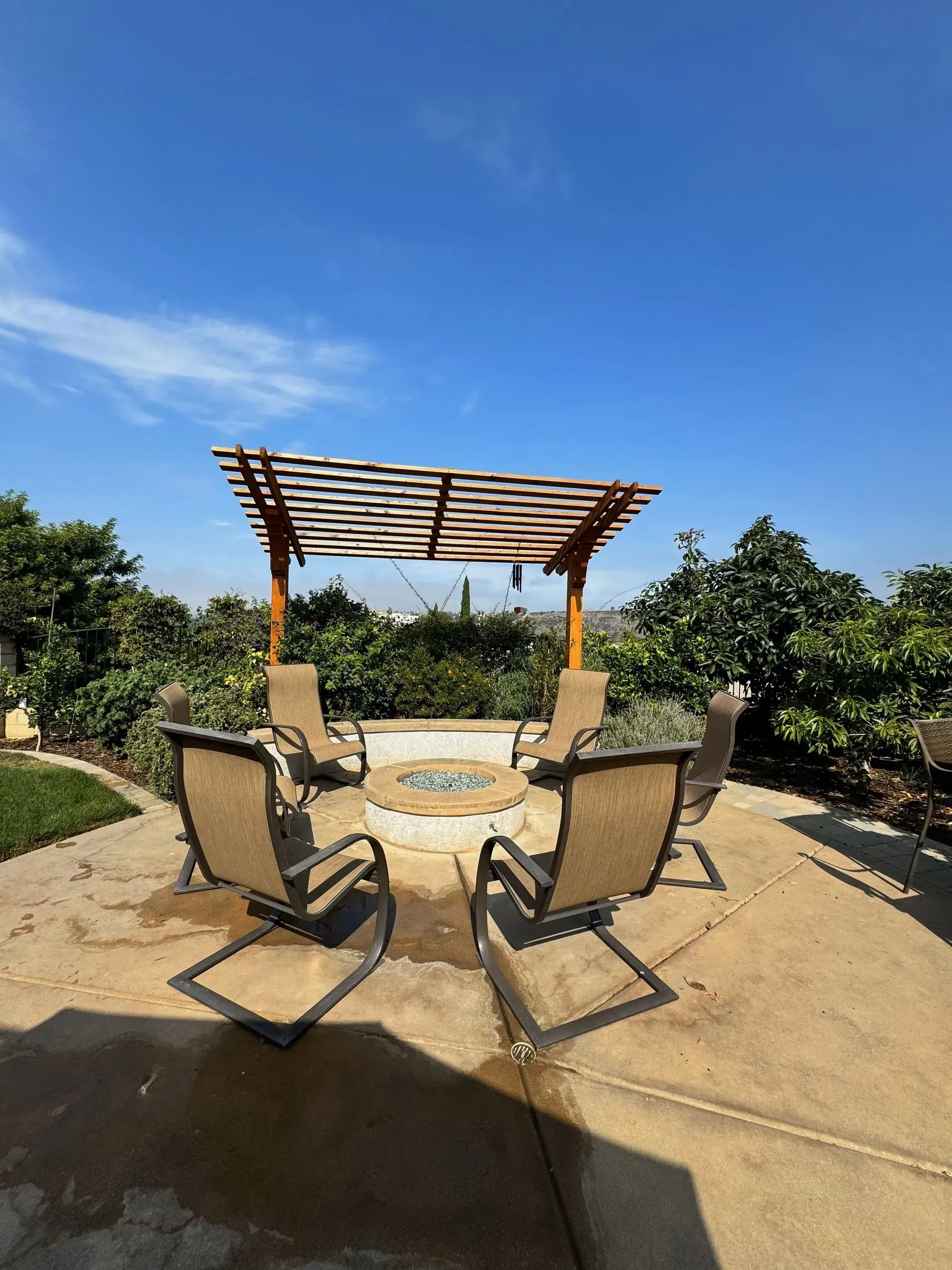 Outdoor fire pit area with chairs and a wooden pergola, under a blue sky.