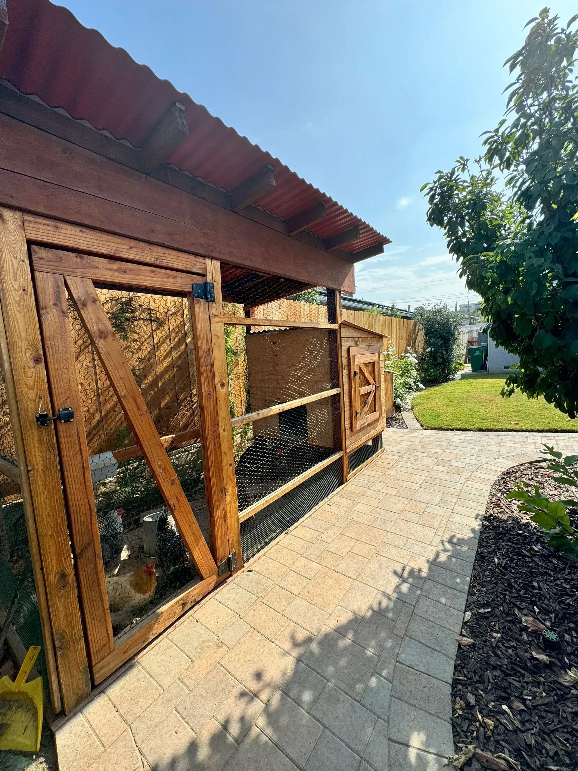 Chicken coop with wooden frame and metal roof, on a brick path, sunny day.