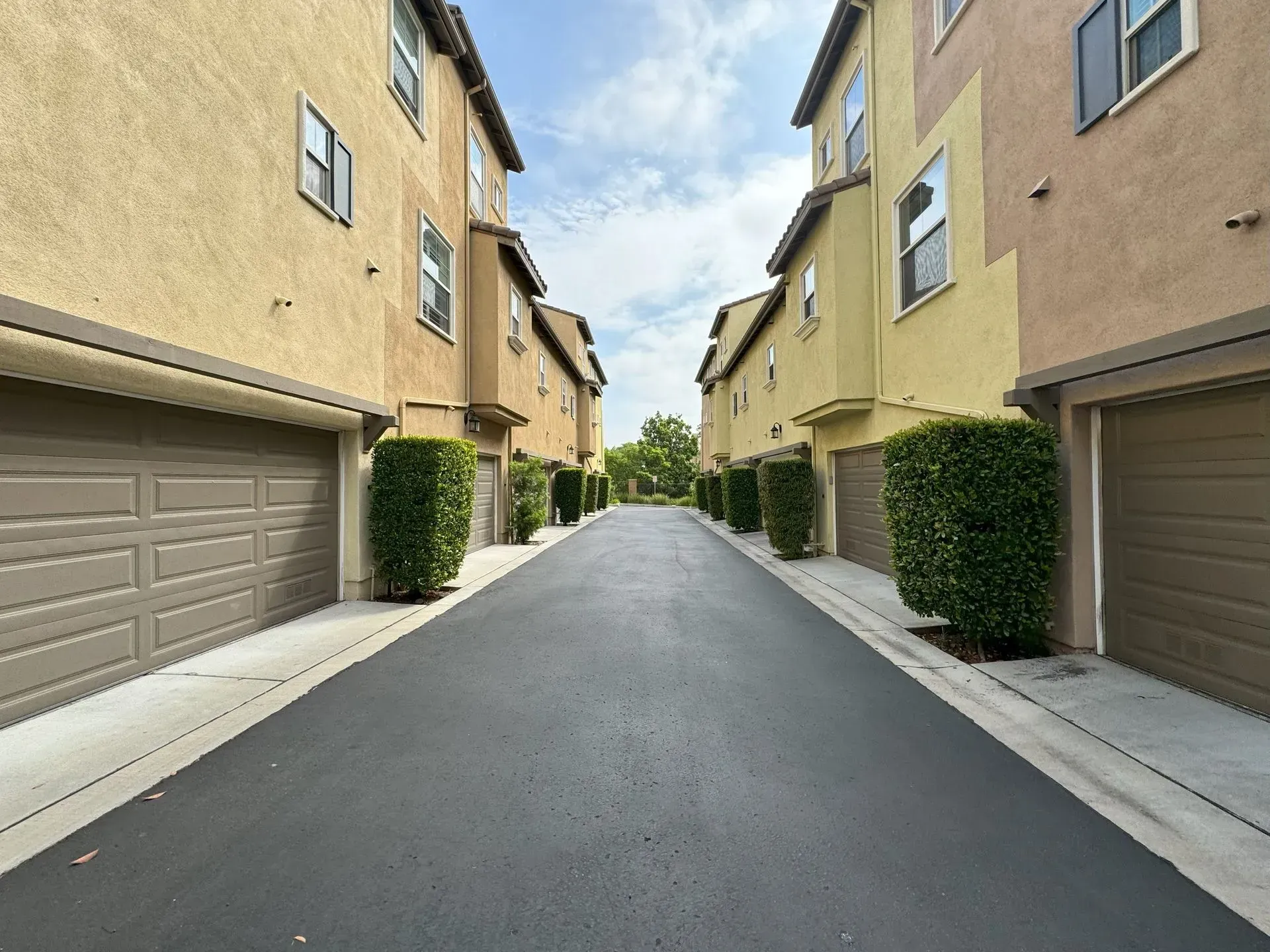 Alleyway between two beige townhouses with garages. Asphalt road and green bushes. Cloudy sky.