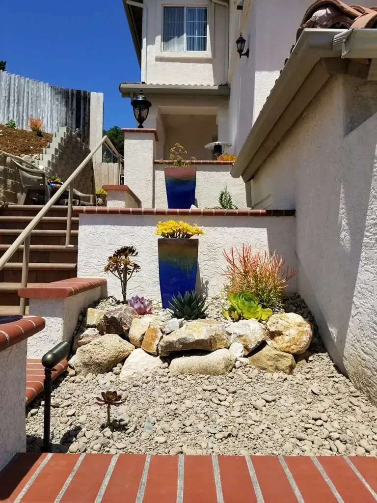 Exterior view of a house with stone steps, gravel, and tiered landscaping. Bright blue sky.