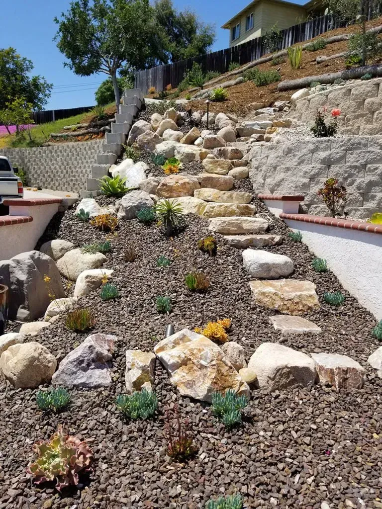 Stone staircase and landscaped hillside with rocks, plants, and retaining walls. Sunny day.