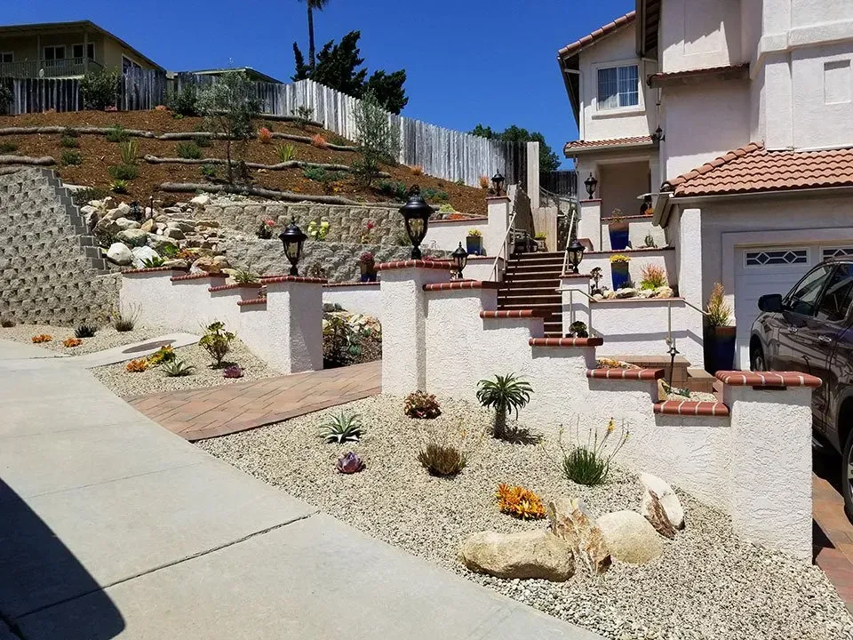 Multi-tiered landscaped yard with stone steps, gravel, retaining walls, and planted areas in front of a house.